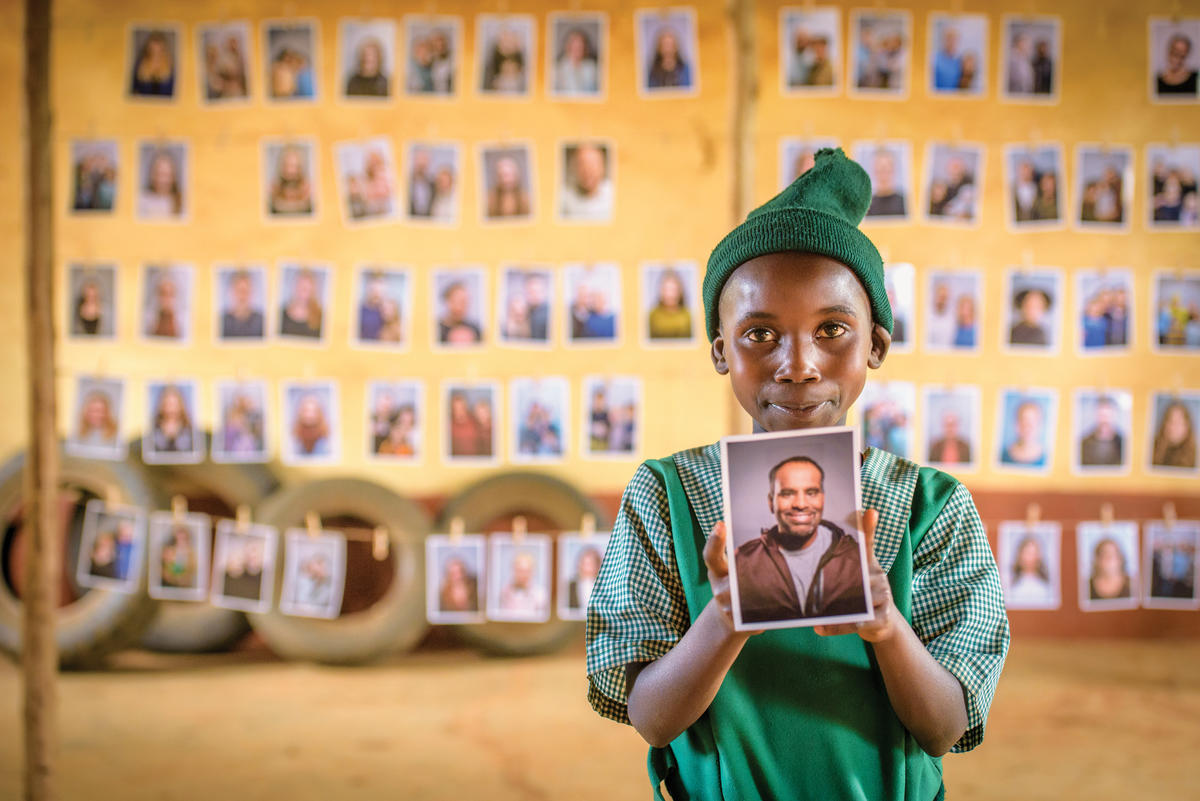 Rosemary from Kenya smiles and holds up a photo of the sponsor she has chosen. There are rows of photos of other sponsors in the background behind her