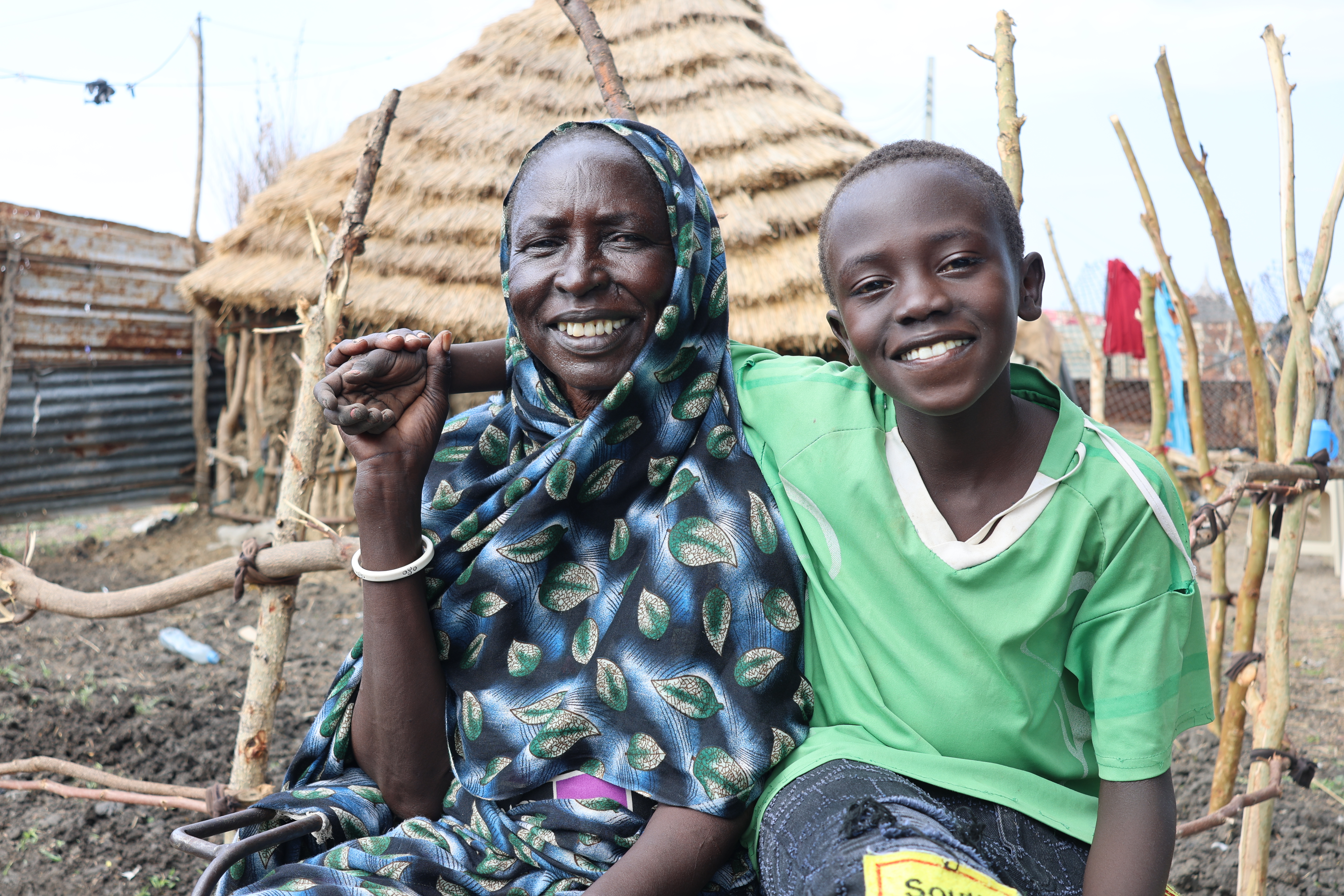 A mother and son, sat in front of a thatched home smiling at the camera, South Sudan.