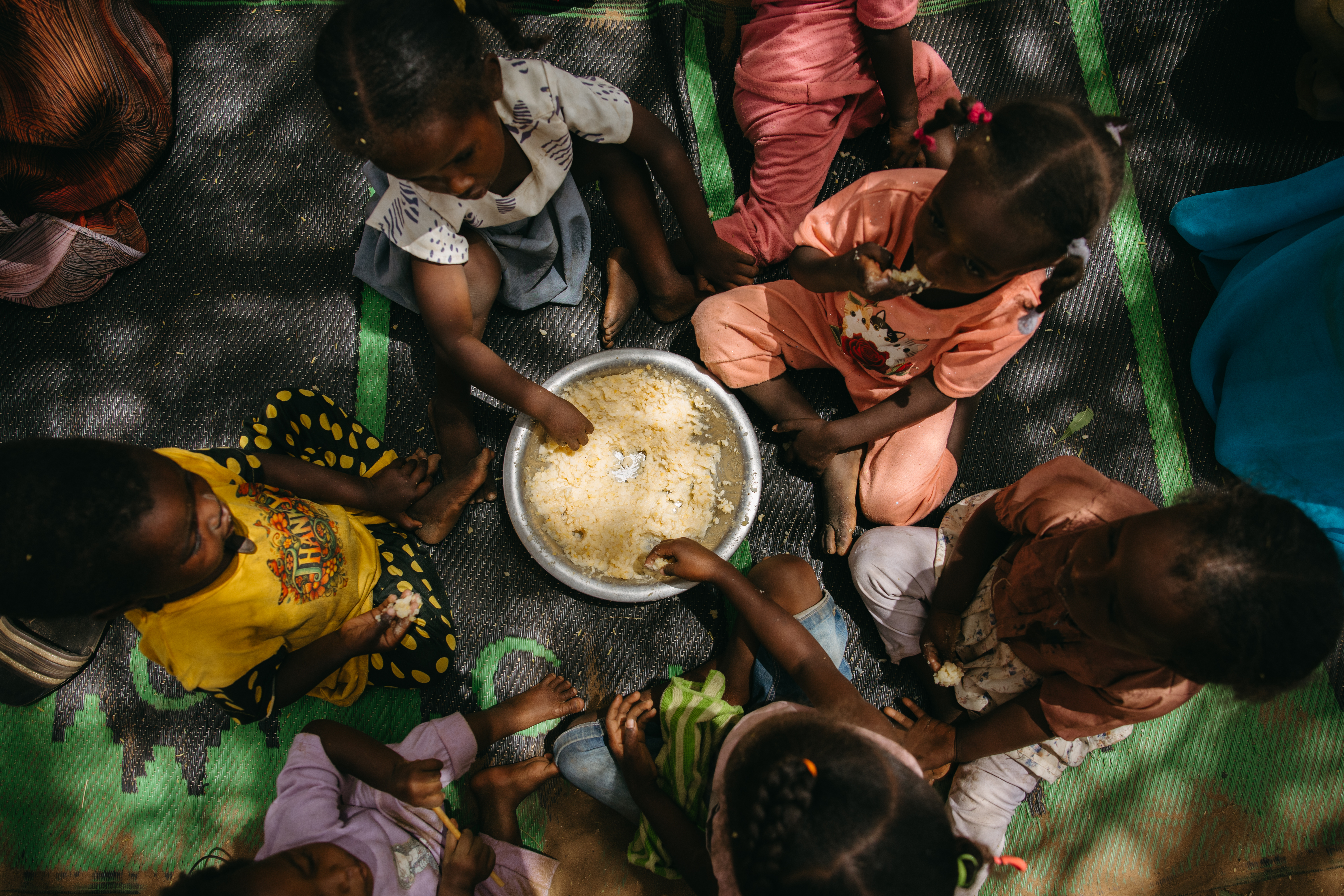 Children sit around a bowl of food taking handfuls. 