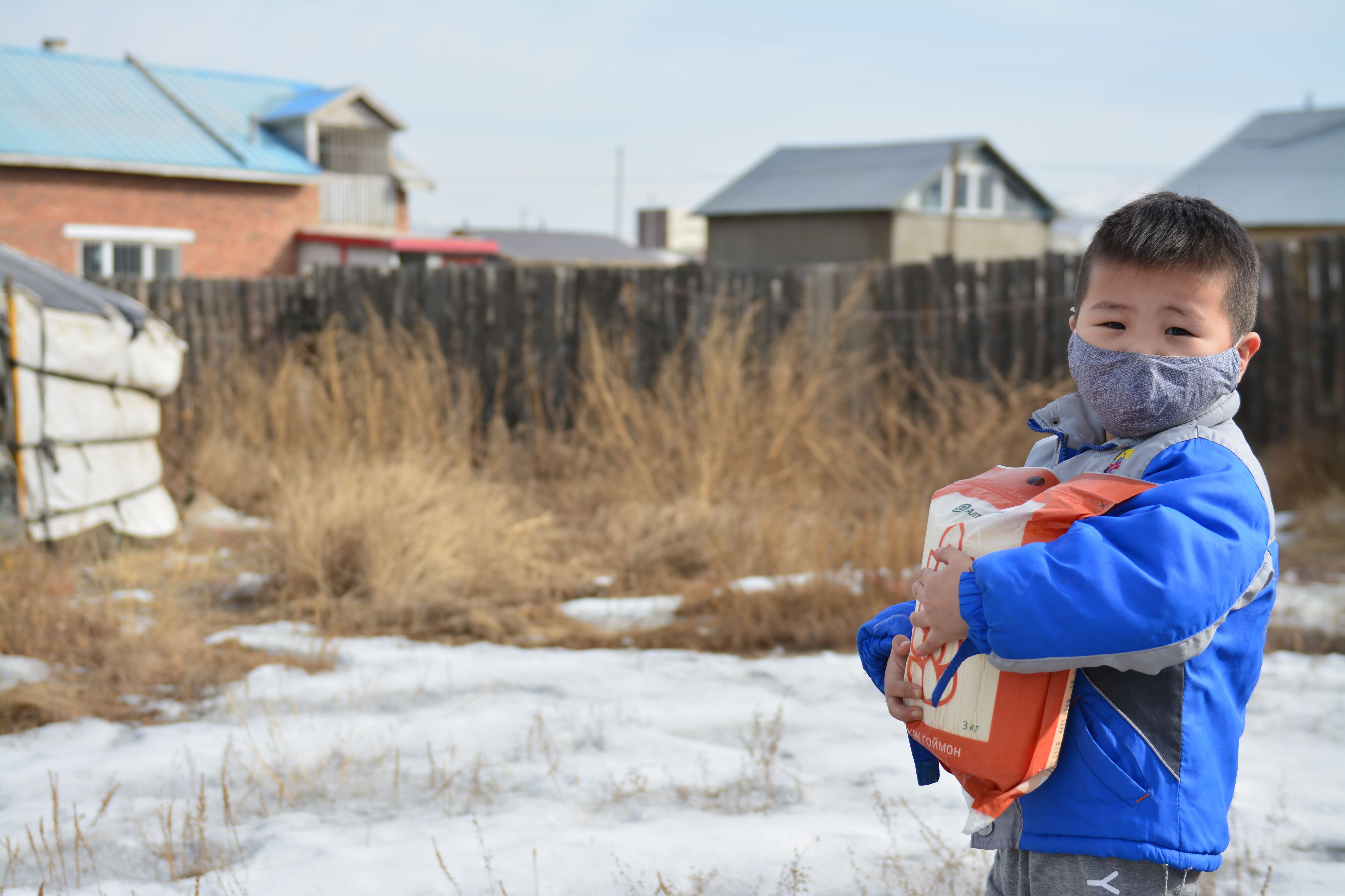 Boy in Mongolia wears a mask and holds a package of food assistance as he stands in a snowy backyard