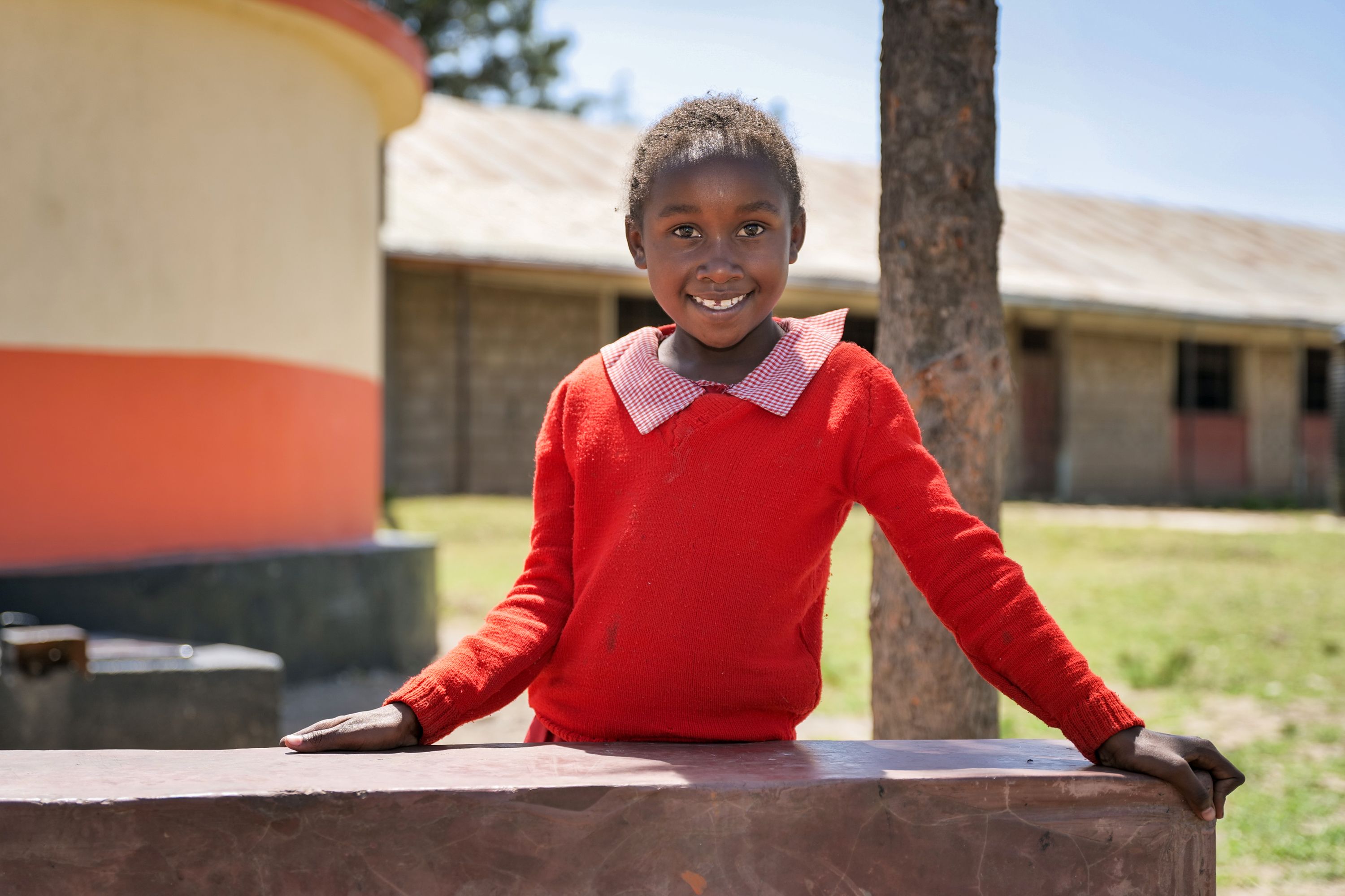 A girl in a red school jumper standing outdoors in front of a school building and tree, looking towards the camera.