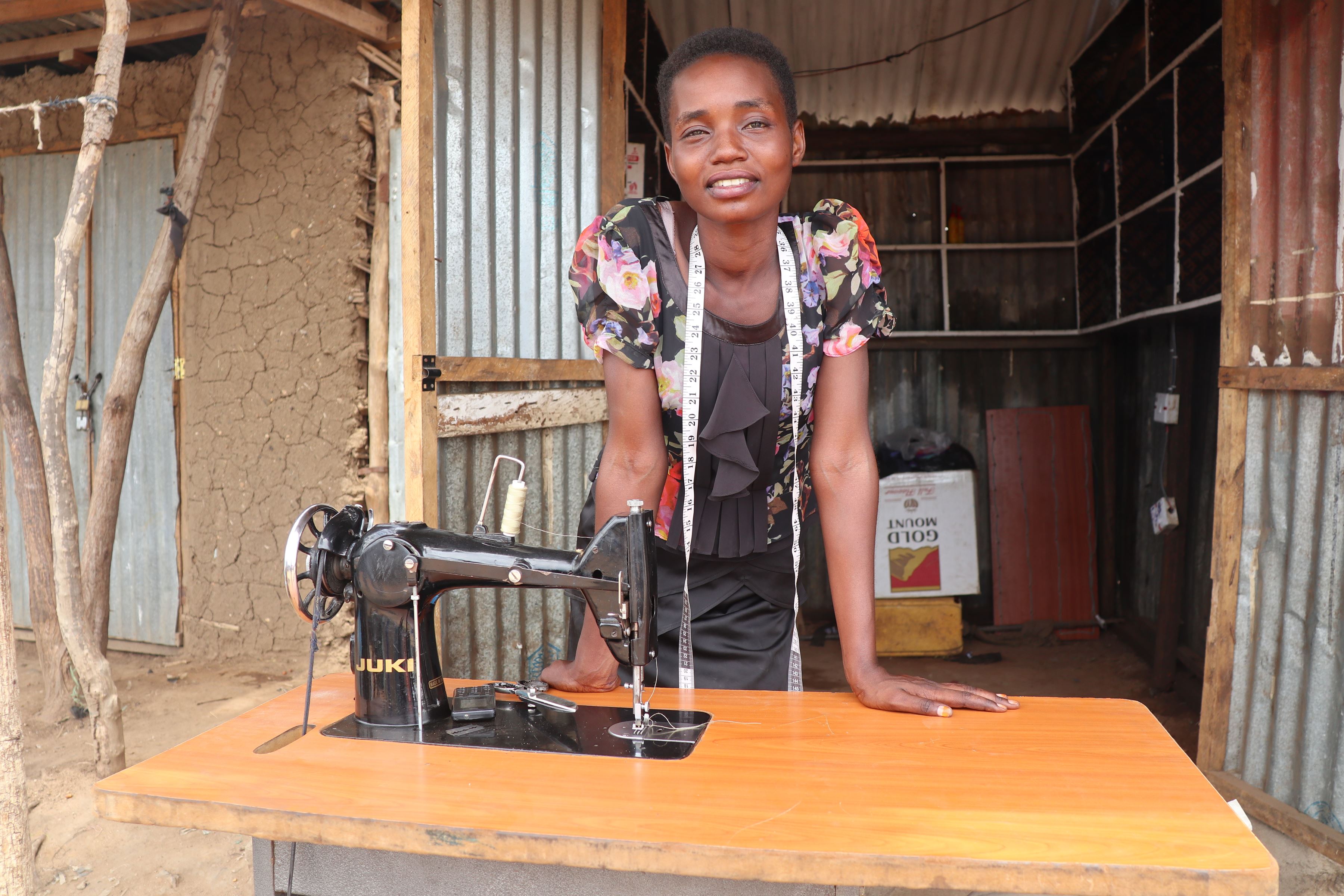 Woman from South Sudan leans on a table outside, with a sewing machine on the table