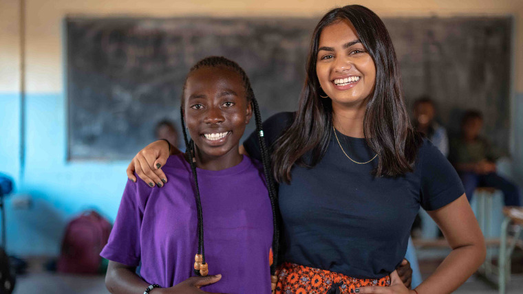 Amika George stands with her arm around a schoolgirl in Zambia, in front of a blackboard