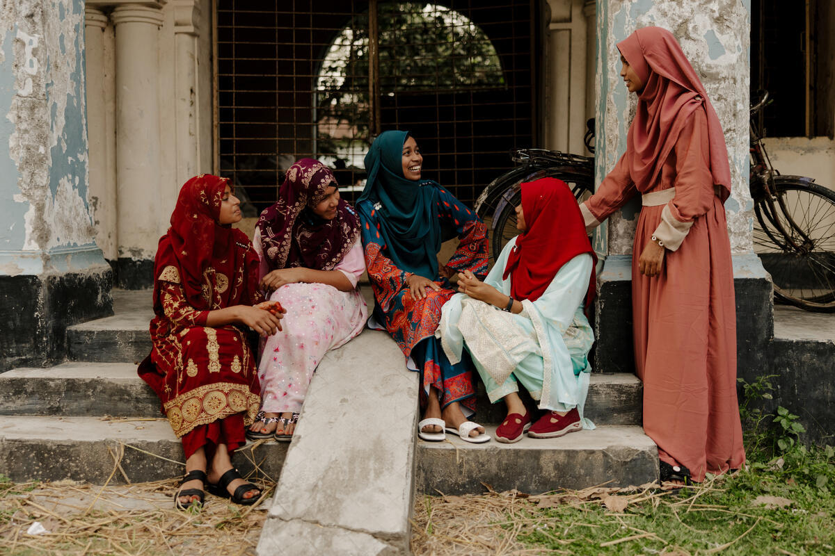 Misu and her friends sit on stone steps in colourful clothes, chatting and laughing together.