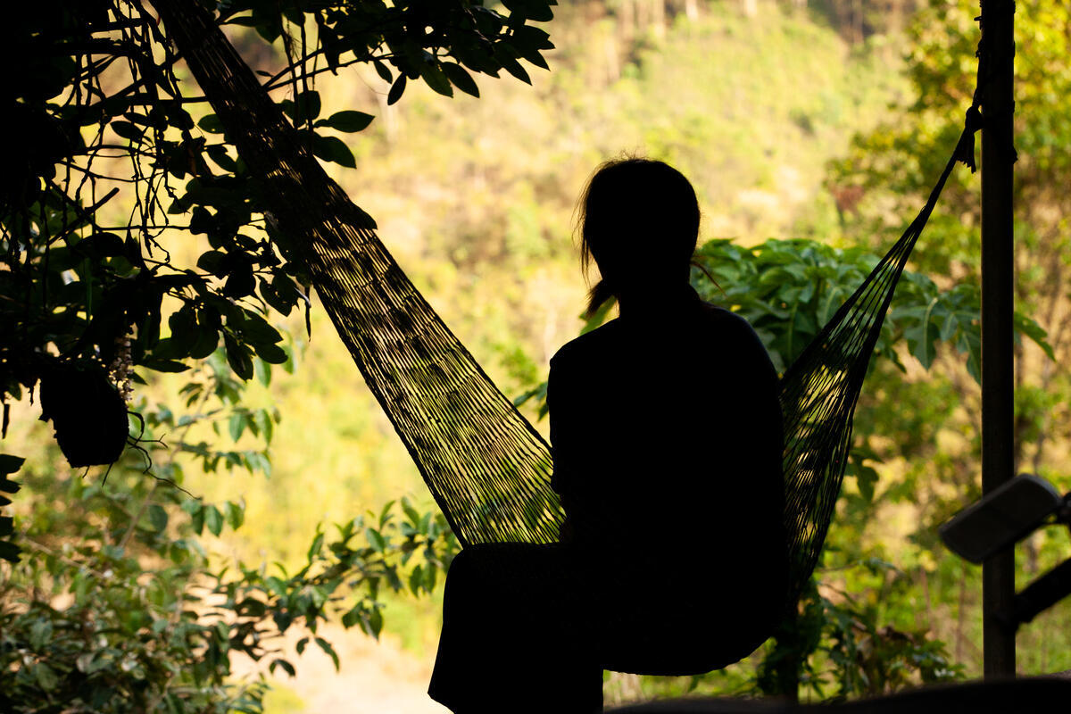 Child sitting in a hammock outdoors surrounded by trees and vegetation.