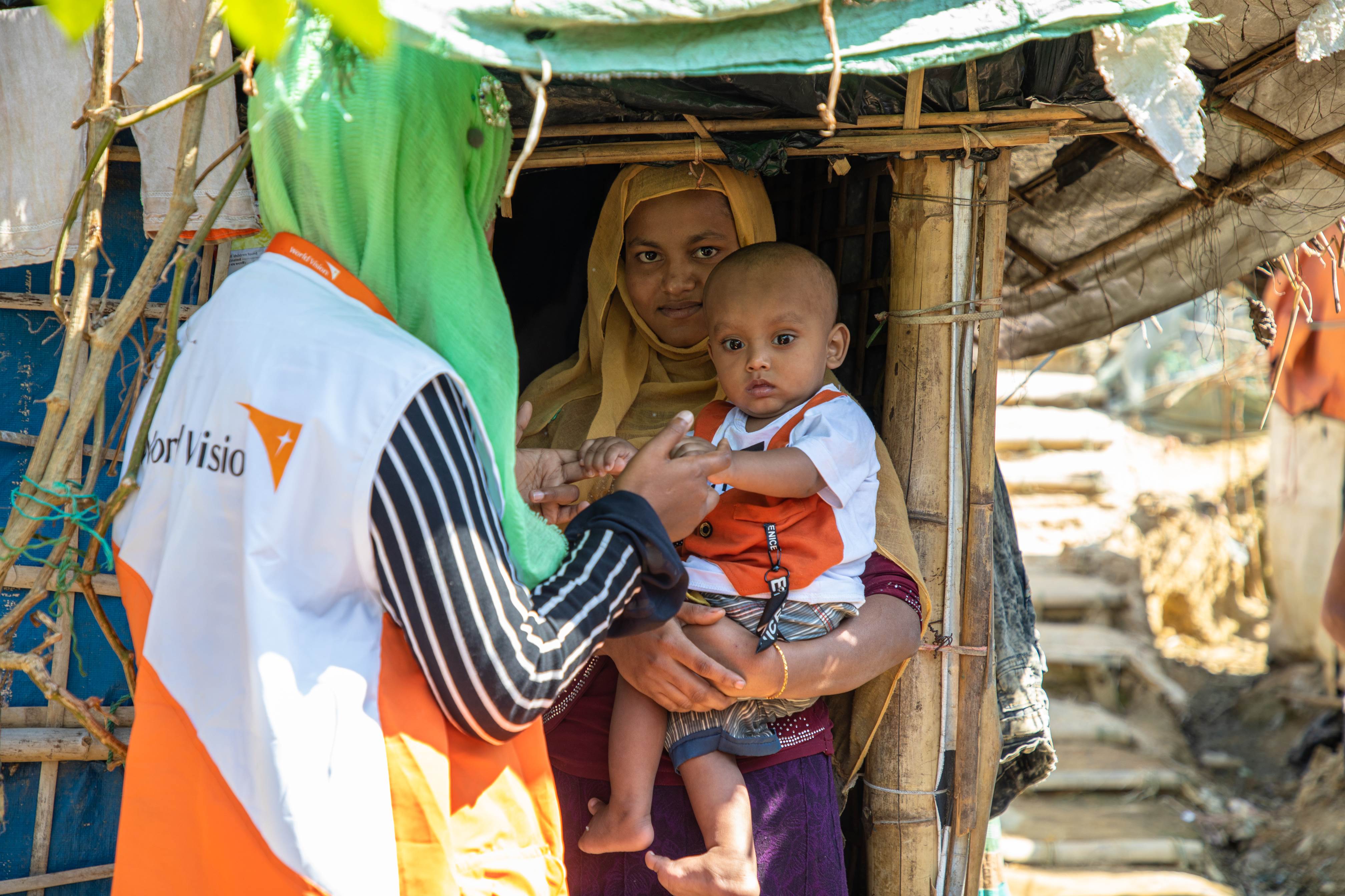 Mother in Bangladesh holding her baby whilst standing in the doorway of her makeshift home talking to a World Vision staff member with back to camera