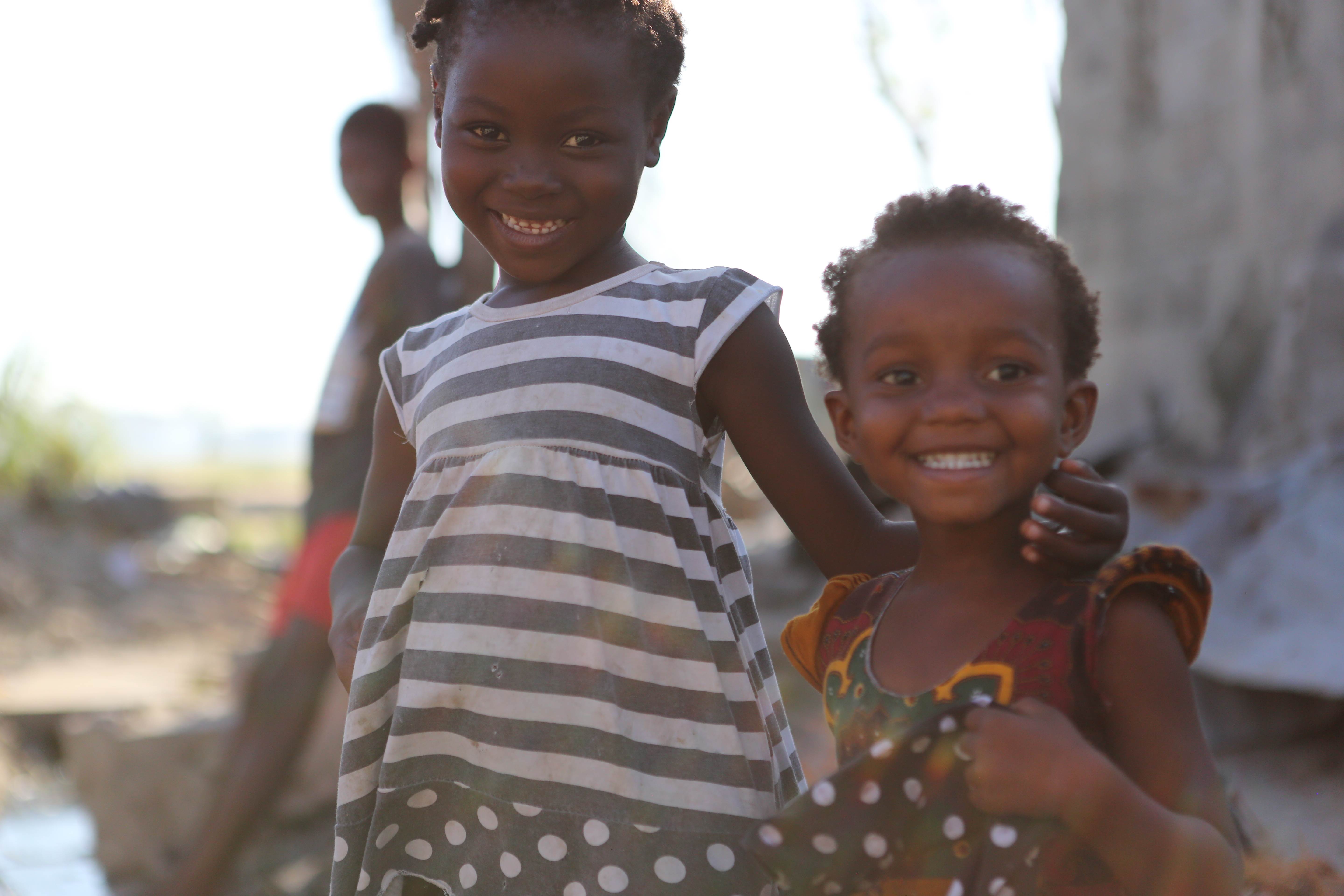 A little girl and her younger sister stand side by side smiling into the camera