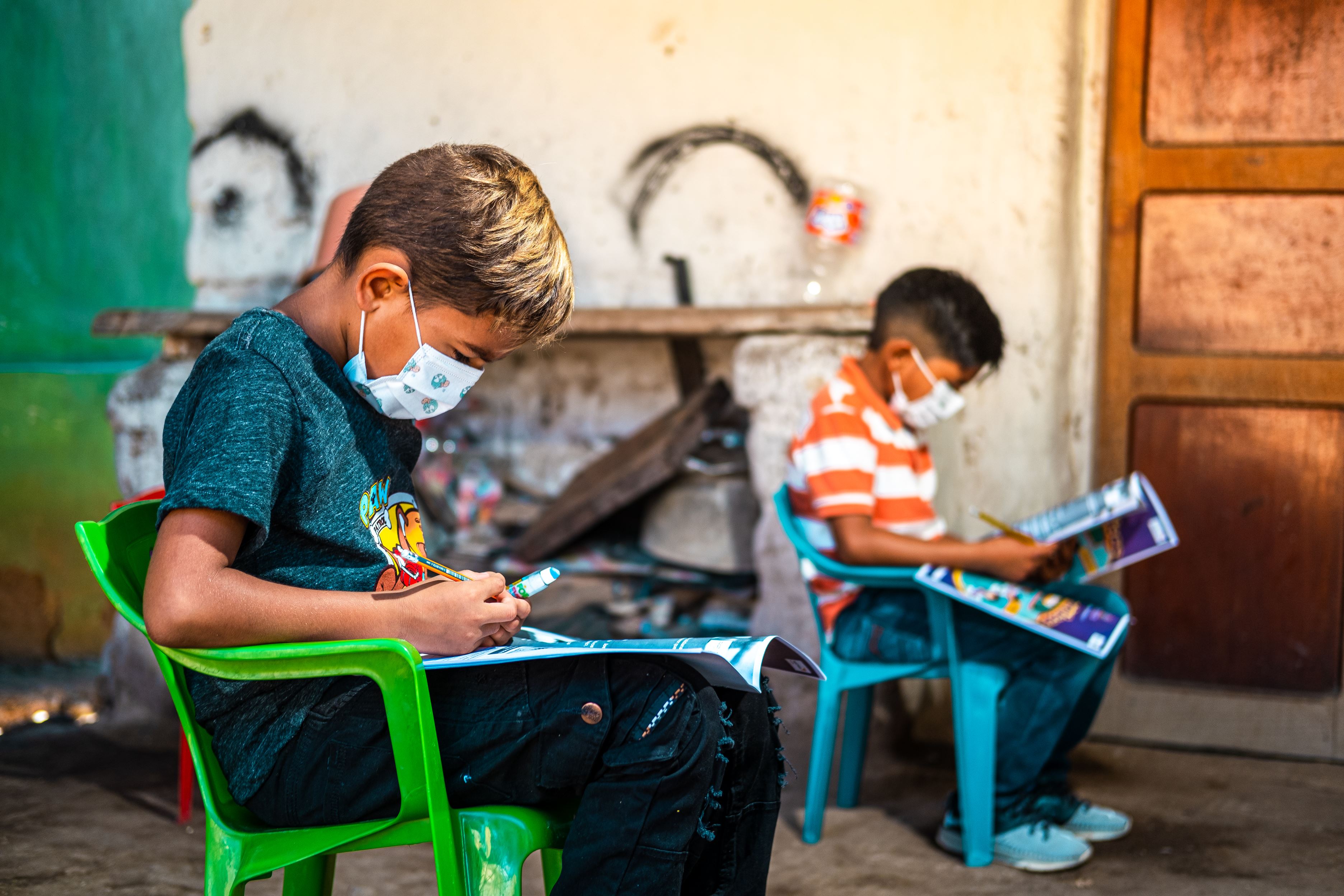 Two children in Honduras sit on plastic chairs and work with masks on due to coronavirus