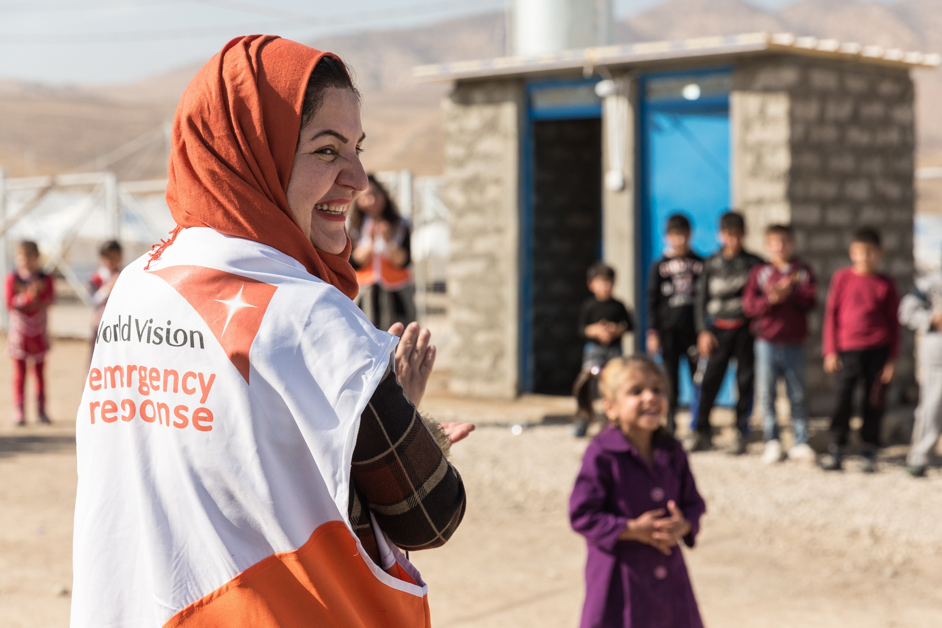 Female World Vision staff member with back to camera, and head turned smiling, with children in the background at a child friendly space in a camp in Iraq