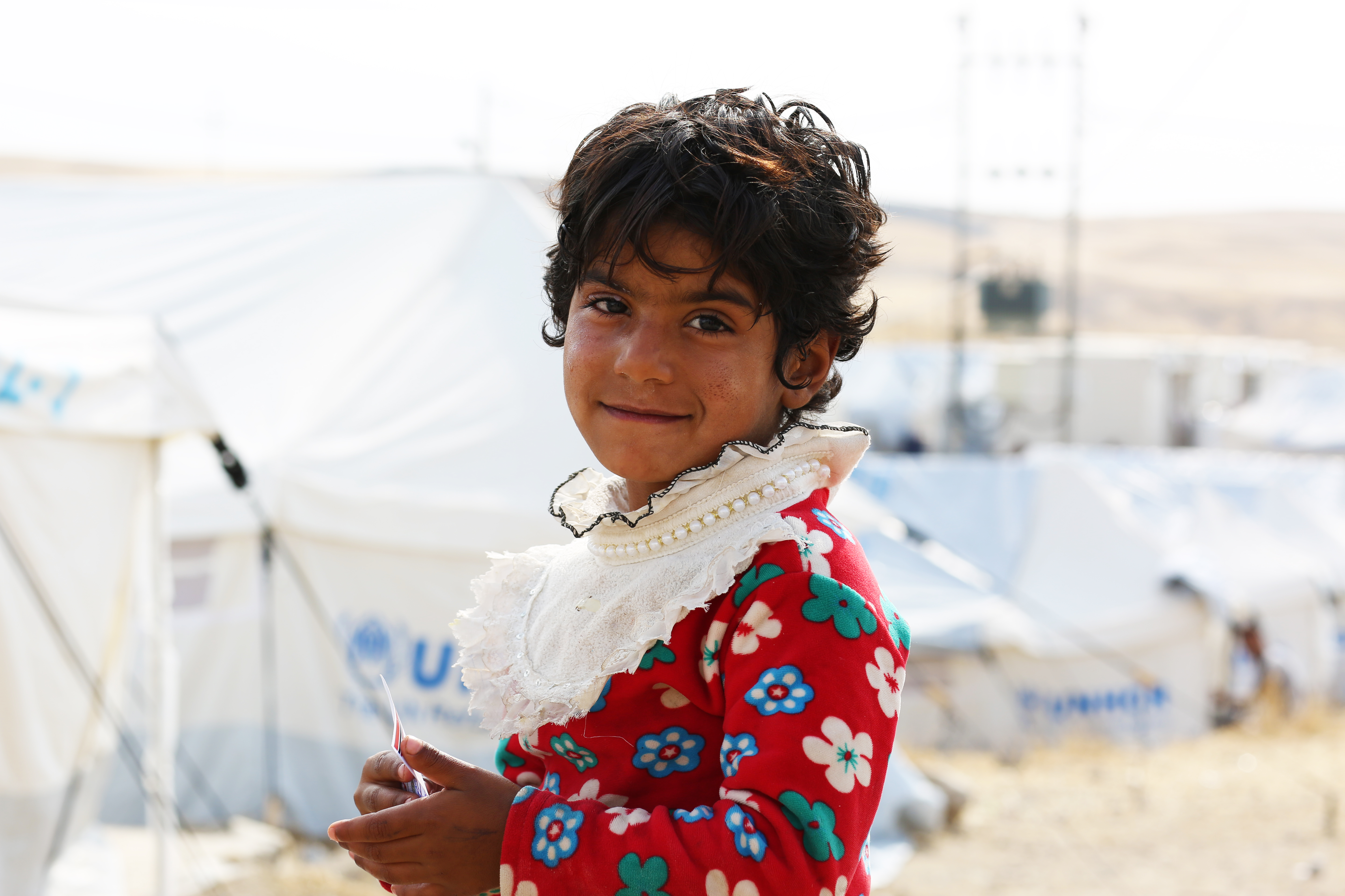 Young Syrian girl in a refugee camp smiles as she stands in front of rows of tents