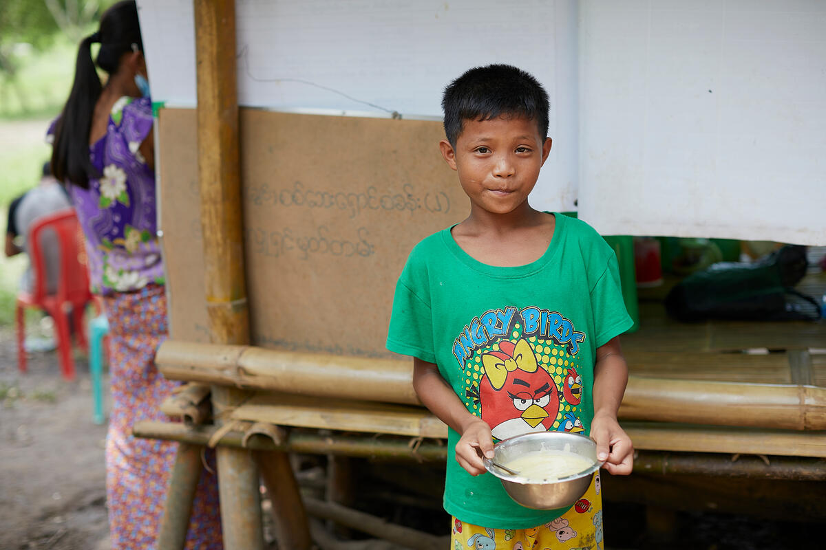 Child from Myanmar carries a pot, standing outside in a green top