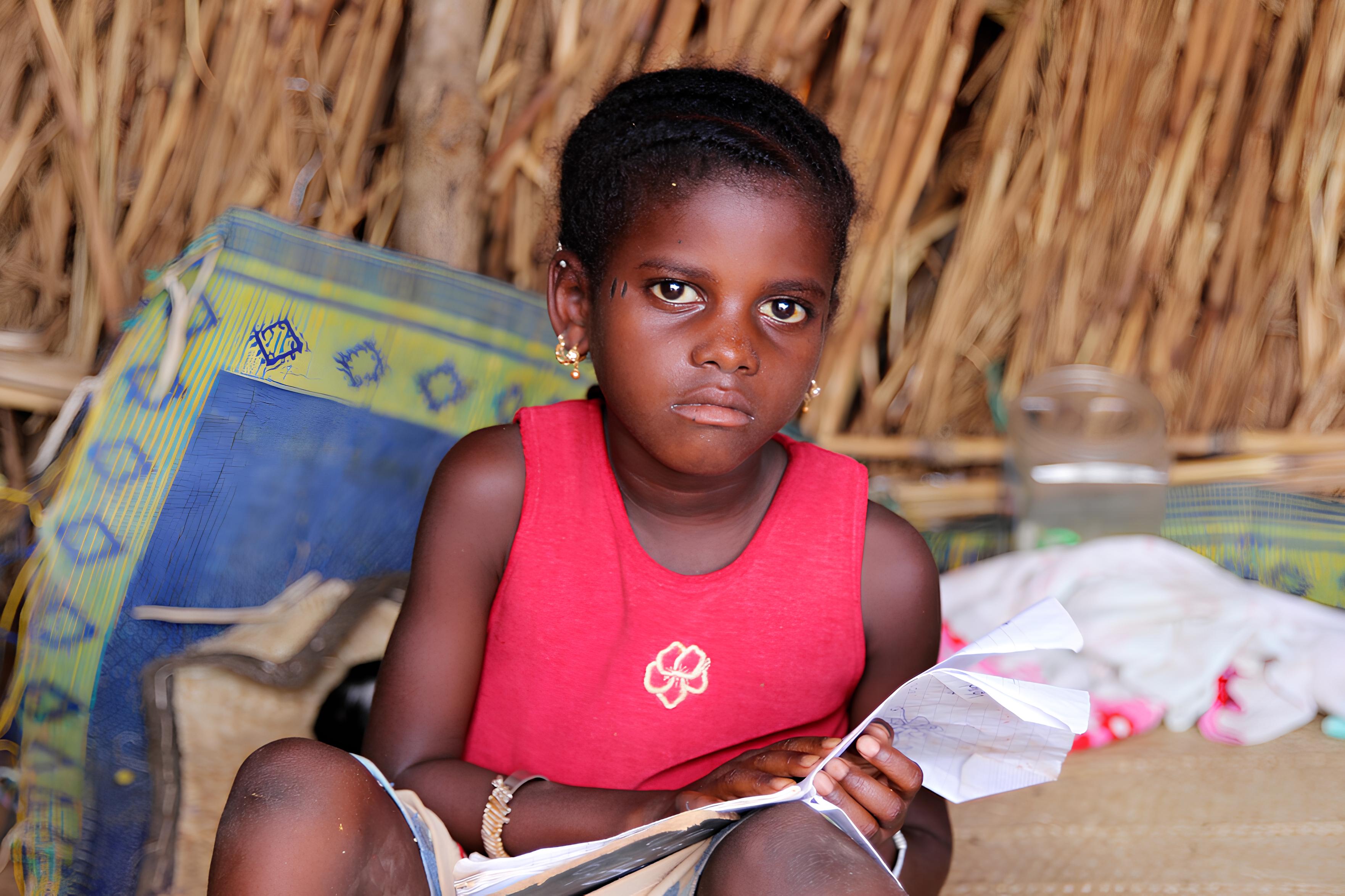 Girl in pink dress sat in front of a thatched backdrop with an open notebook in her hands