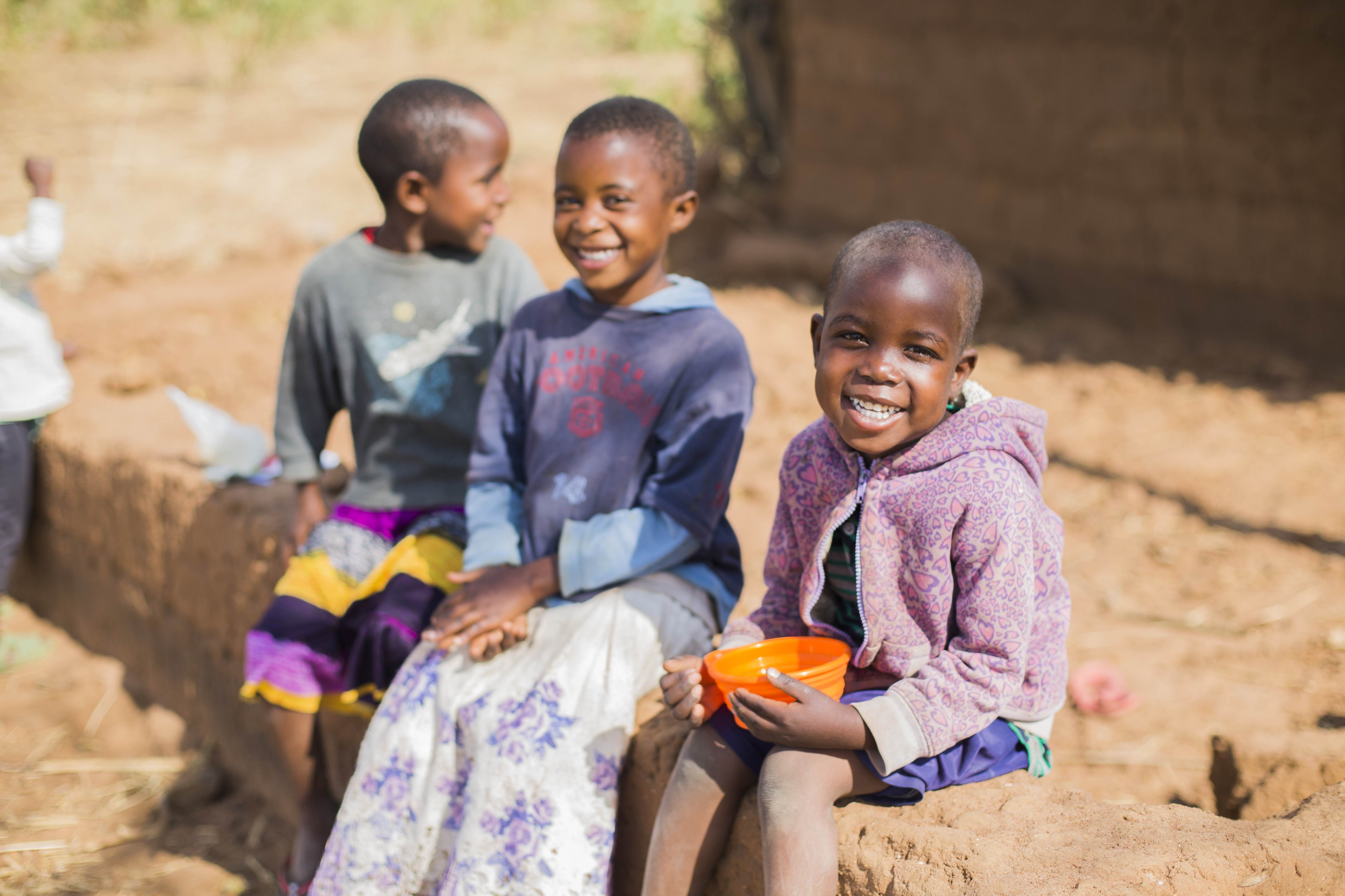 Three children from Tanzania sitting on a mud brick wall with two smiling to the camera