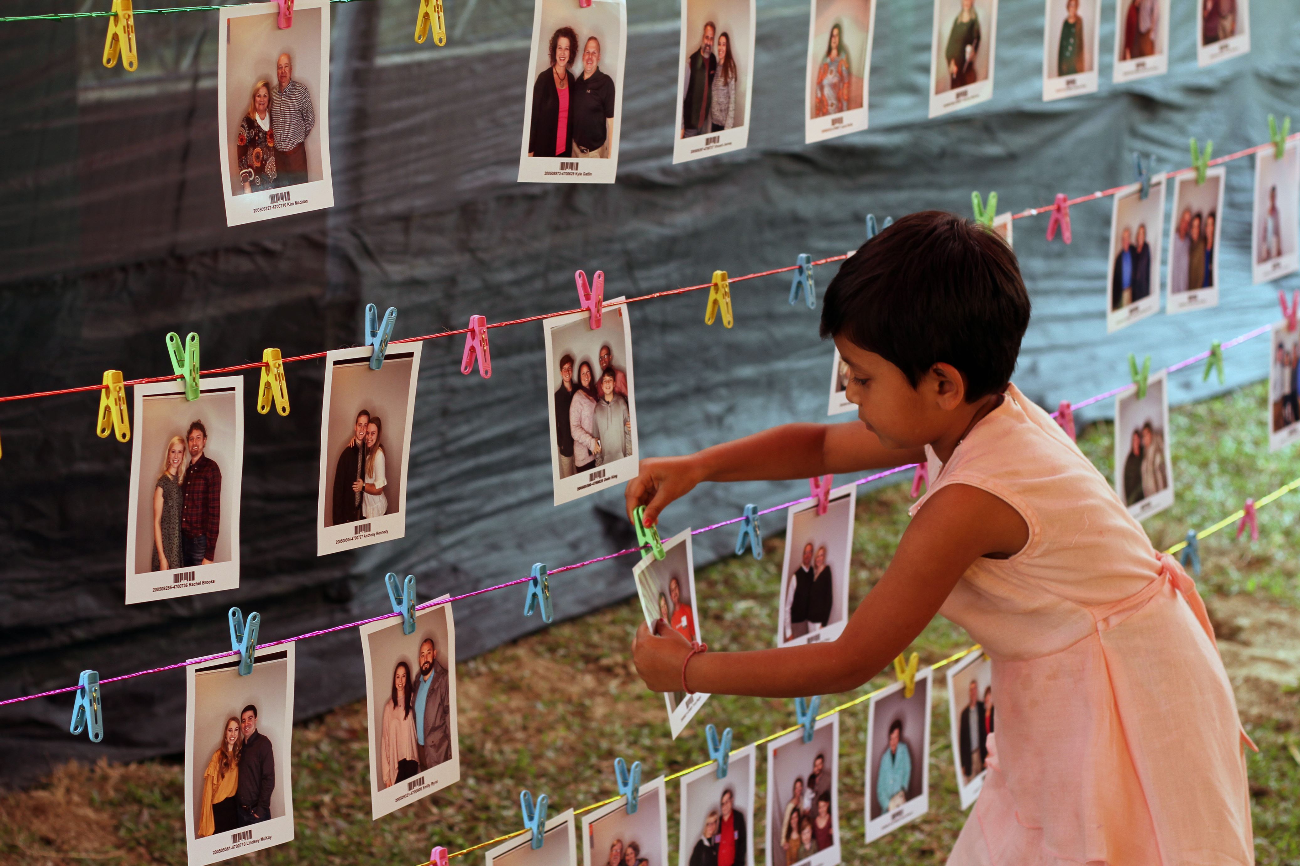 Girl in Bangladesh in a pink dress reaches to pick a photo from three lines worth of potential sponsor photos hanging from wires in a room