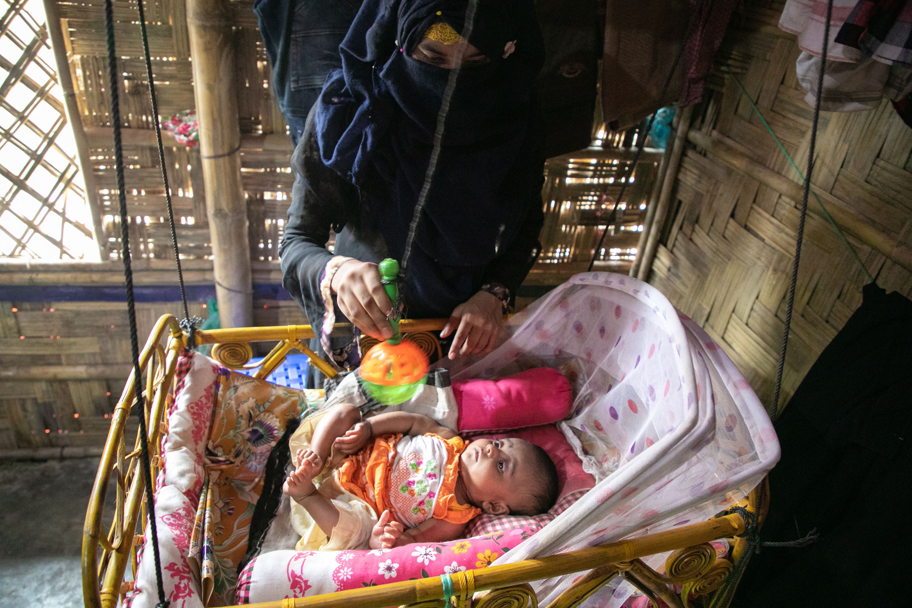 A baby in a cot, while her mother gives her a rattle toy.
