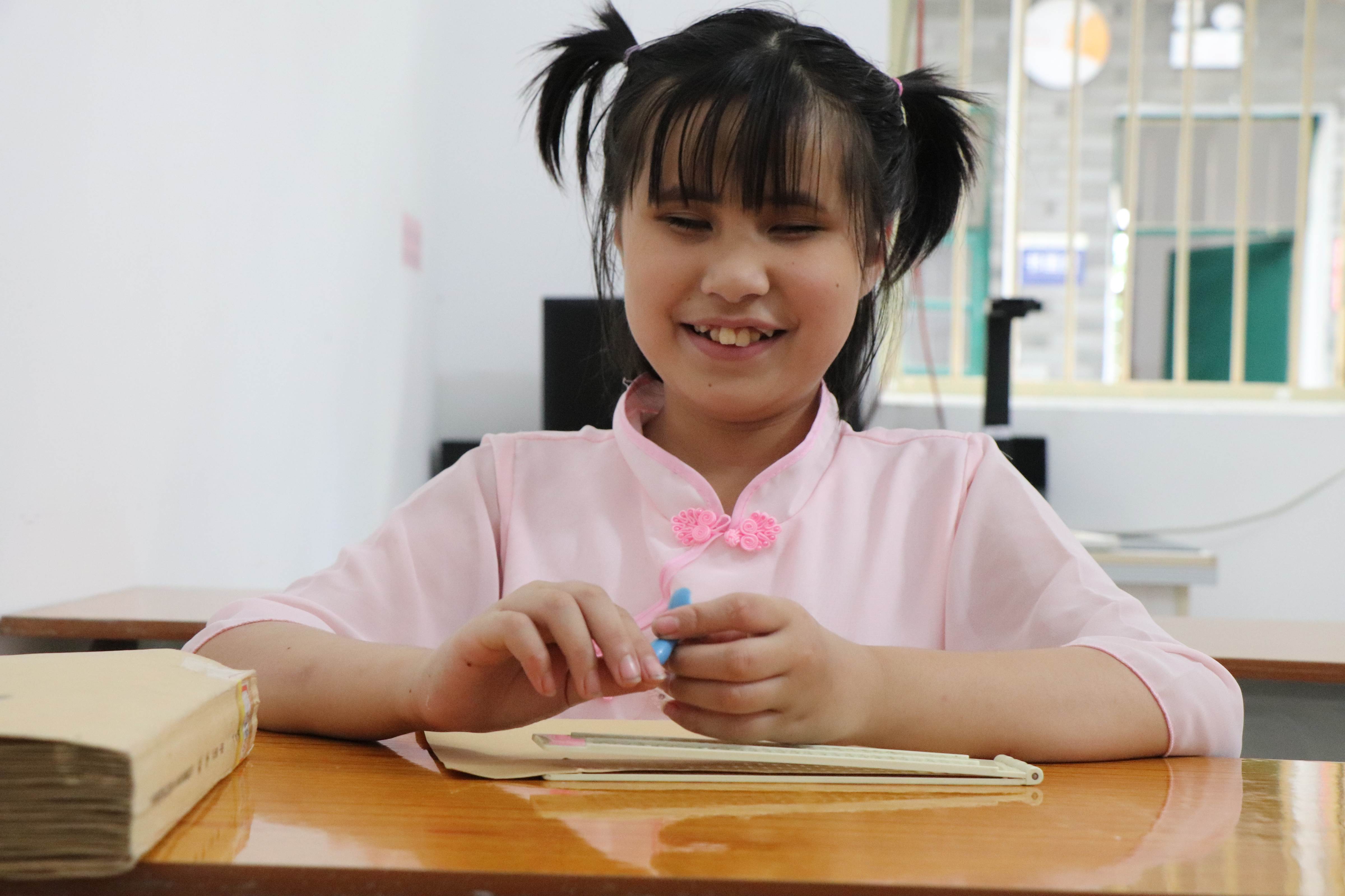 A young girl wearing pink traditional style Chinese blouse, sits at her desk smiling