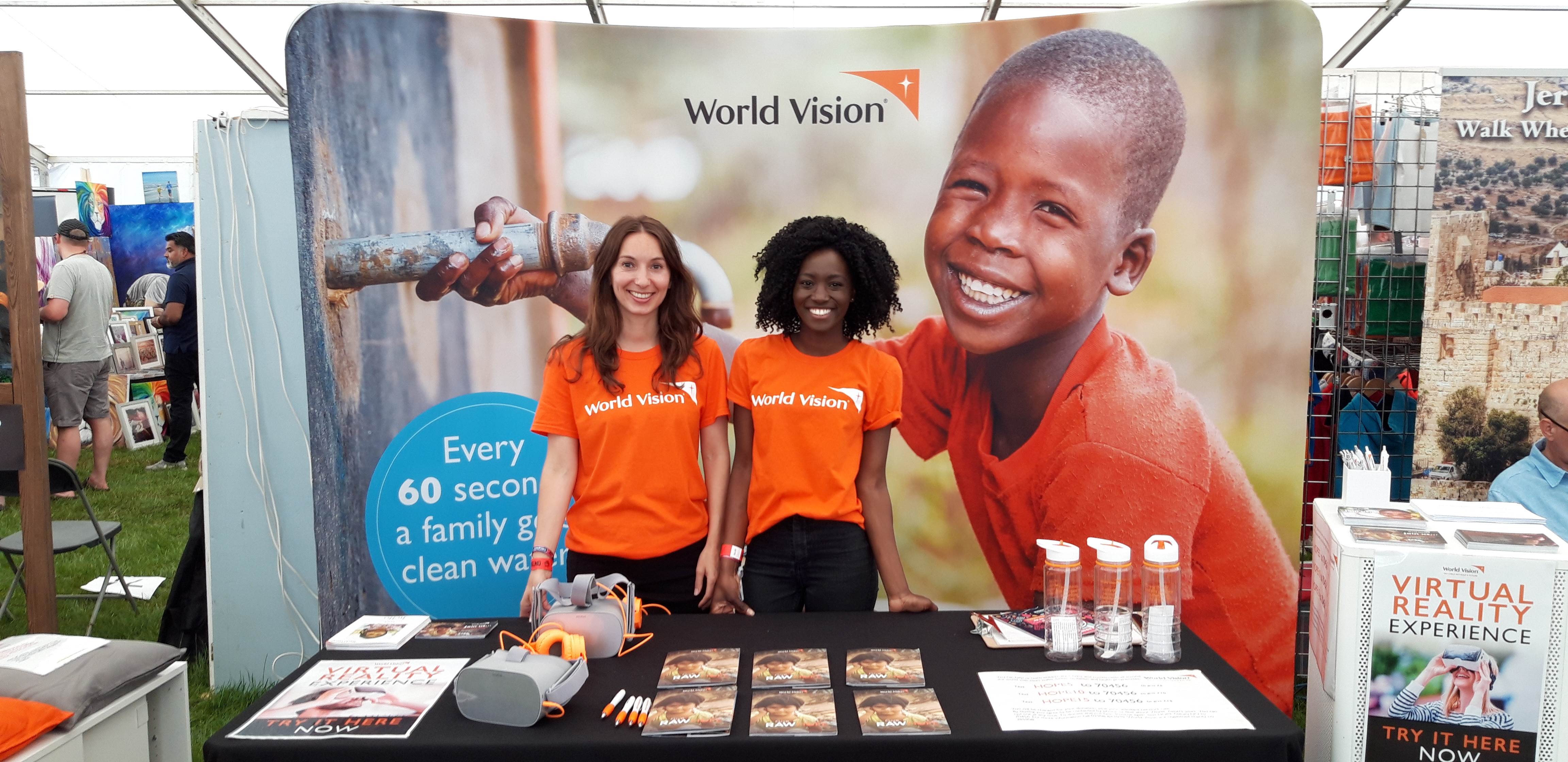 Two women stand at festival stall smiling in front of World Vision banner