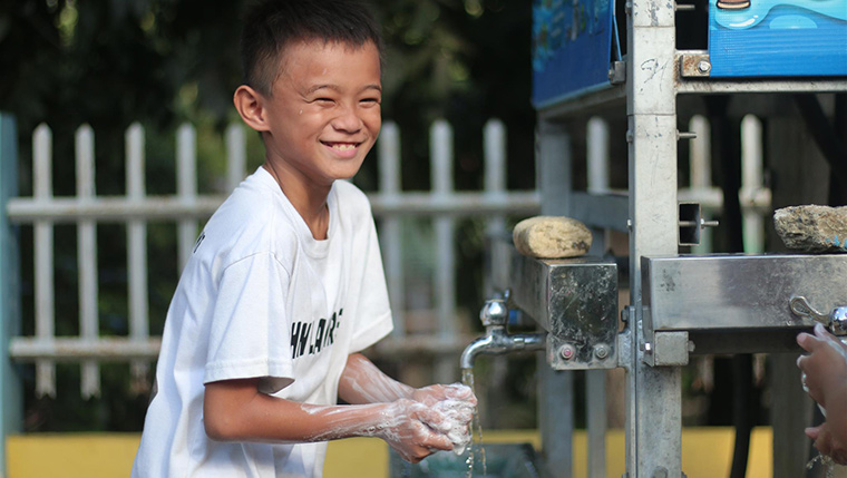A young boy in the Philippines grins as he washes his hands in soap underneath a clean water tap