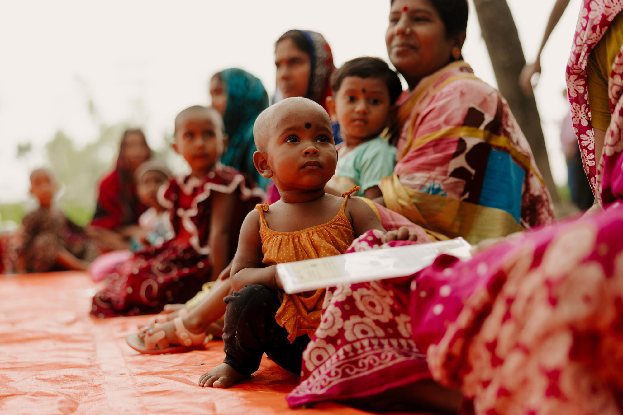 Child sits on a mat alongside community members gathered outdoors in Bangladesh