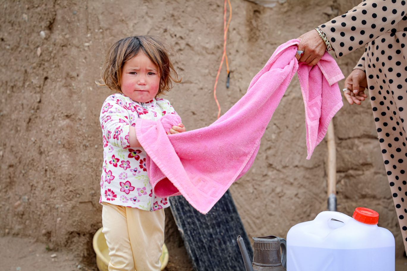 Young refugee child in Afghanistan stands outside, washing their hands with a pink towel and a bottle of water