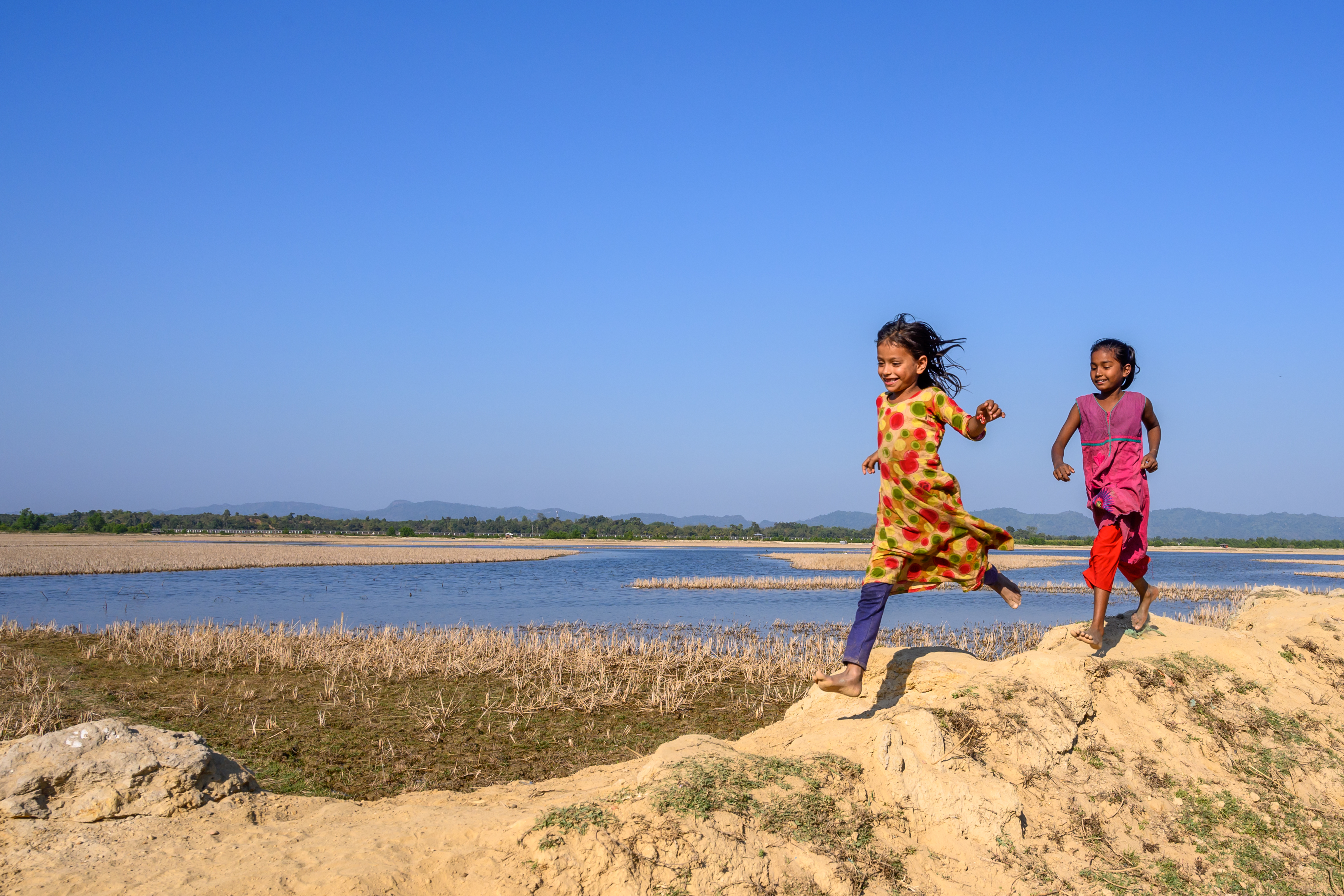 Two girls run along the banks of the Naf River in Bangladesh. Across the water, below clear blue skies in their former home, Myanmar