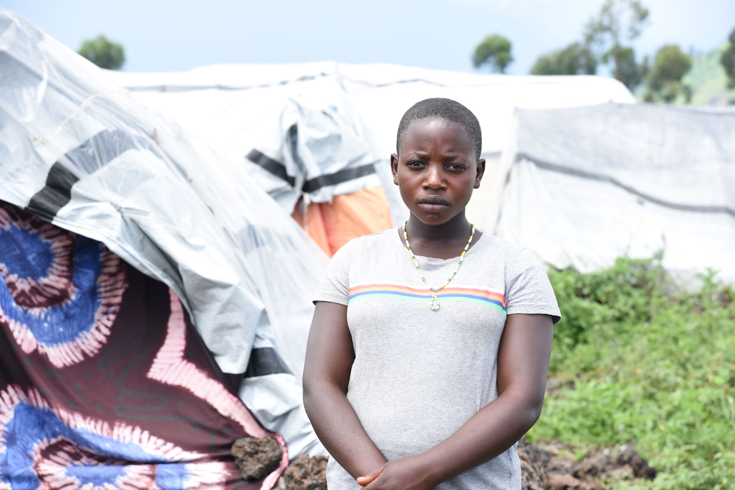 A young girl from the Democratic Republic of Congo looks thoughtful in a white t-shirt, standing in front of a  displacement camp tent