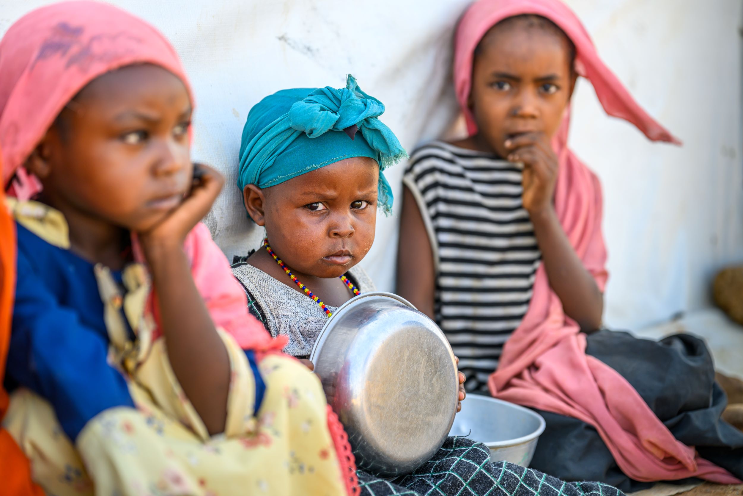 Three young girls from Sudan sit on the floor holding metal bowls