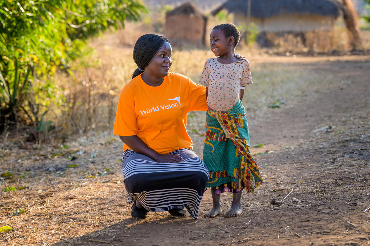World Vision community development worker in an orange branded tshirt crouches down and smiles and a young girl who is smiling back at her in Malawi