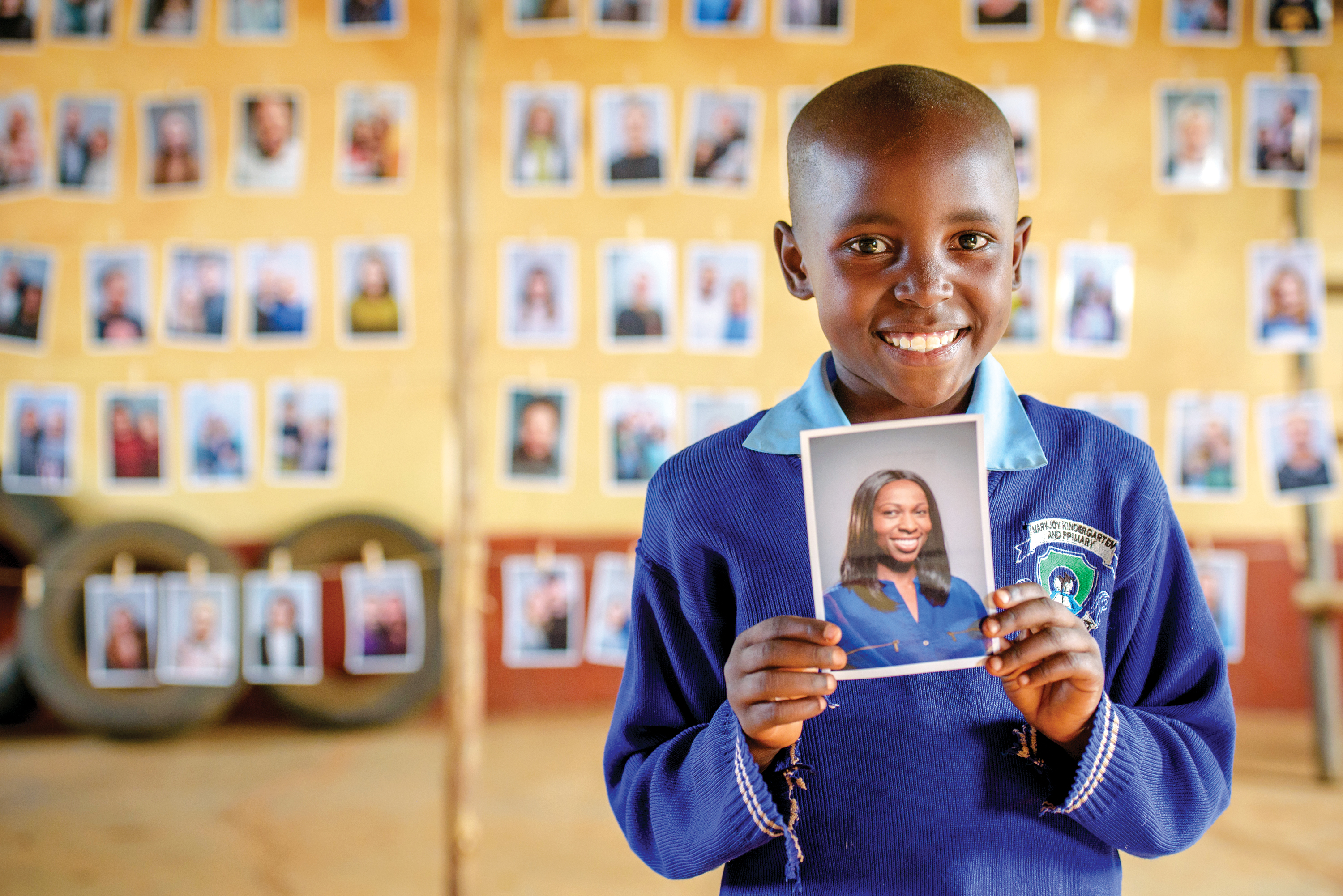 Mary from Kenya smiles and holds up a photo of the sponsor she has chosen. There are rows of photos of other sponsors in the background behind her