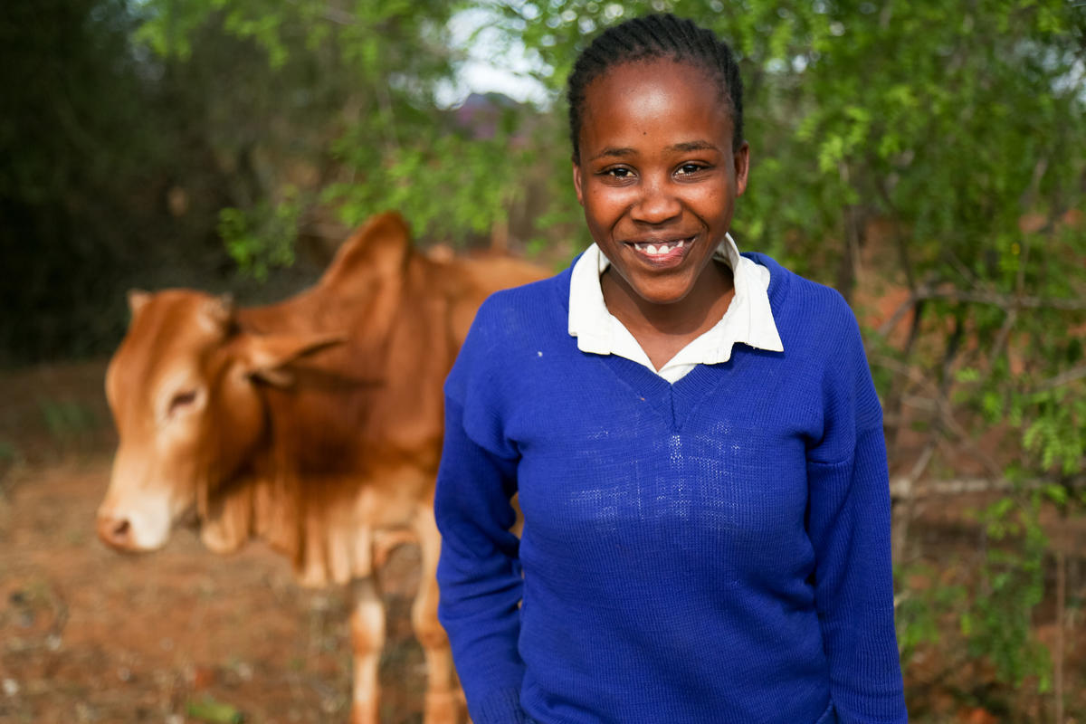 Girl in Kenya smiles with school uniform on and cow behind her