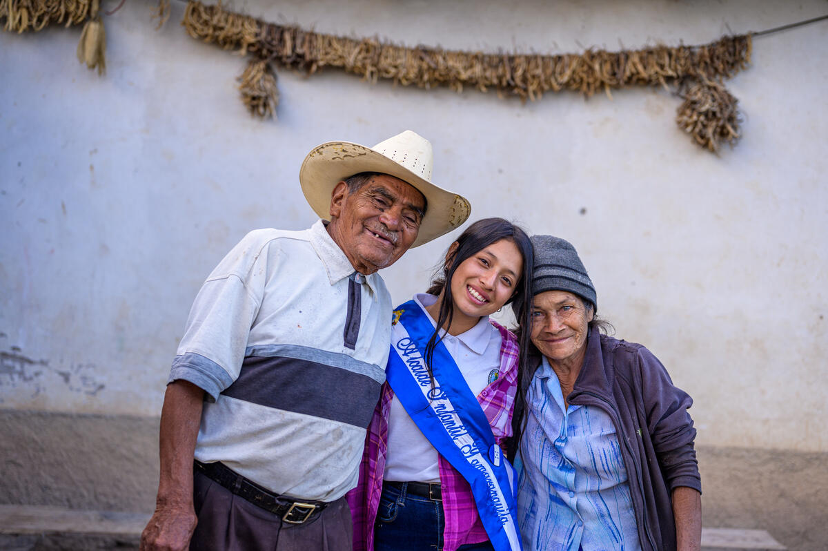 13 years old girl from Honduras is hugging her grandparents, wearing a blue sash signifying she is the youth mayor of her area