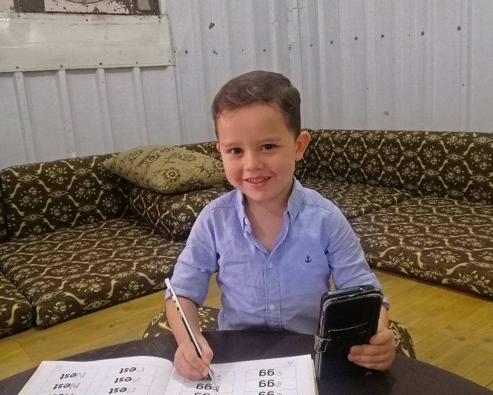 Young boy sits on the floor smiling to the camera with a workbook open and a pencil in his hand