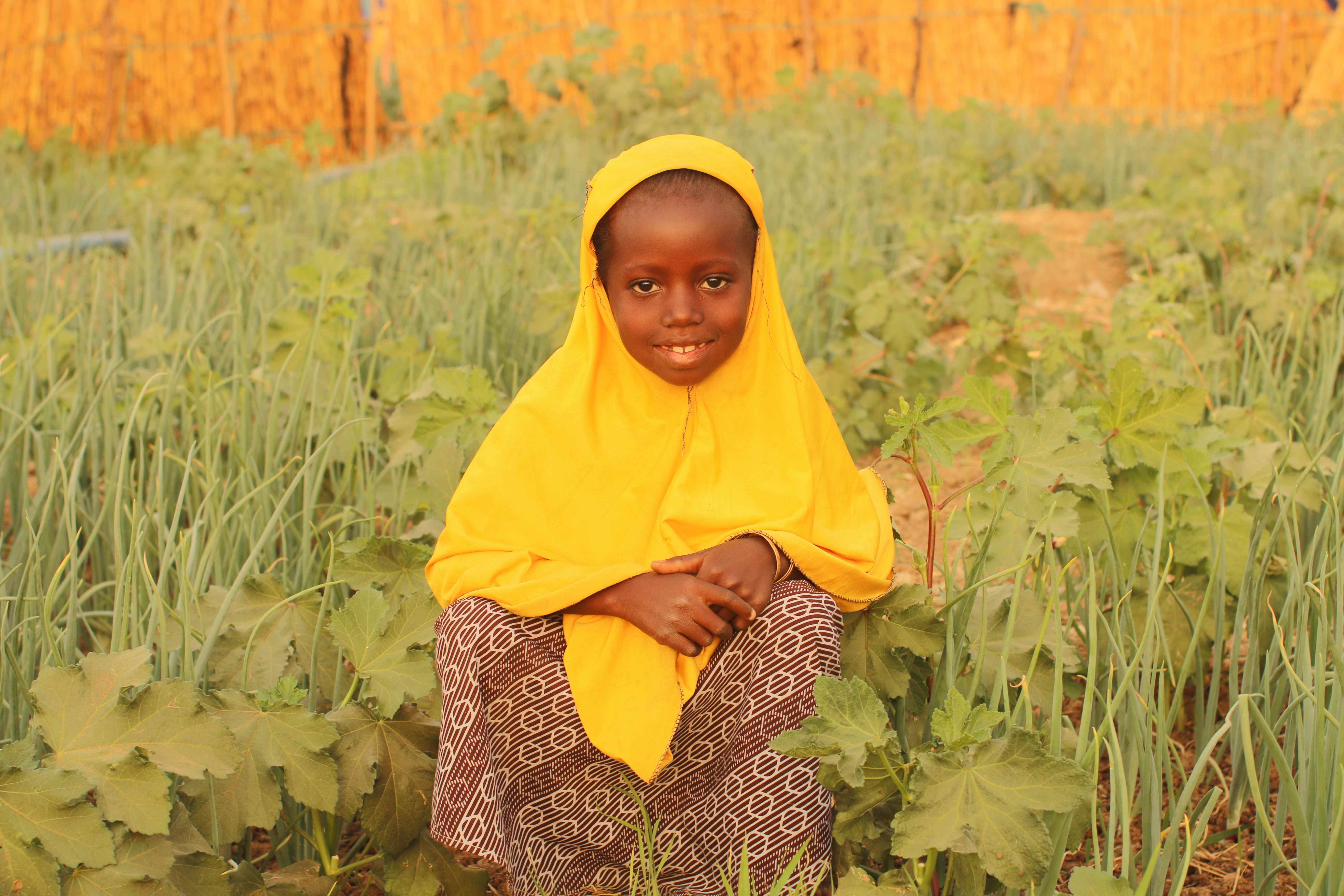 Child from Niger stands in a field of cauliflower, wearing a vibrant yellow headdress