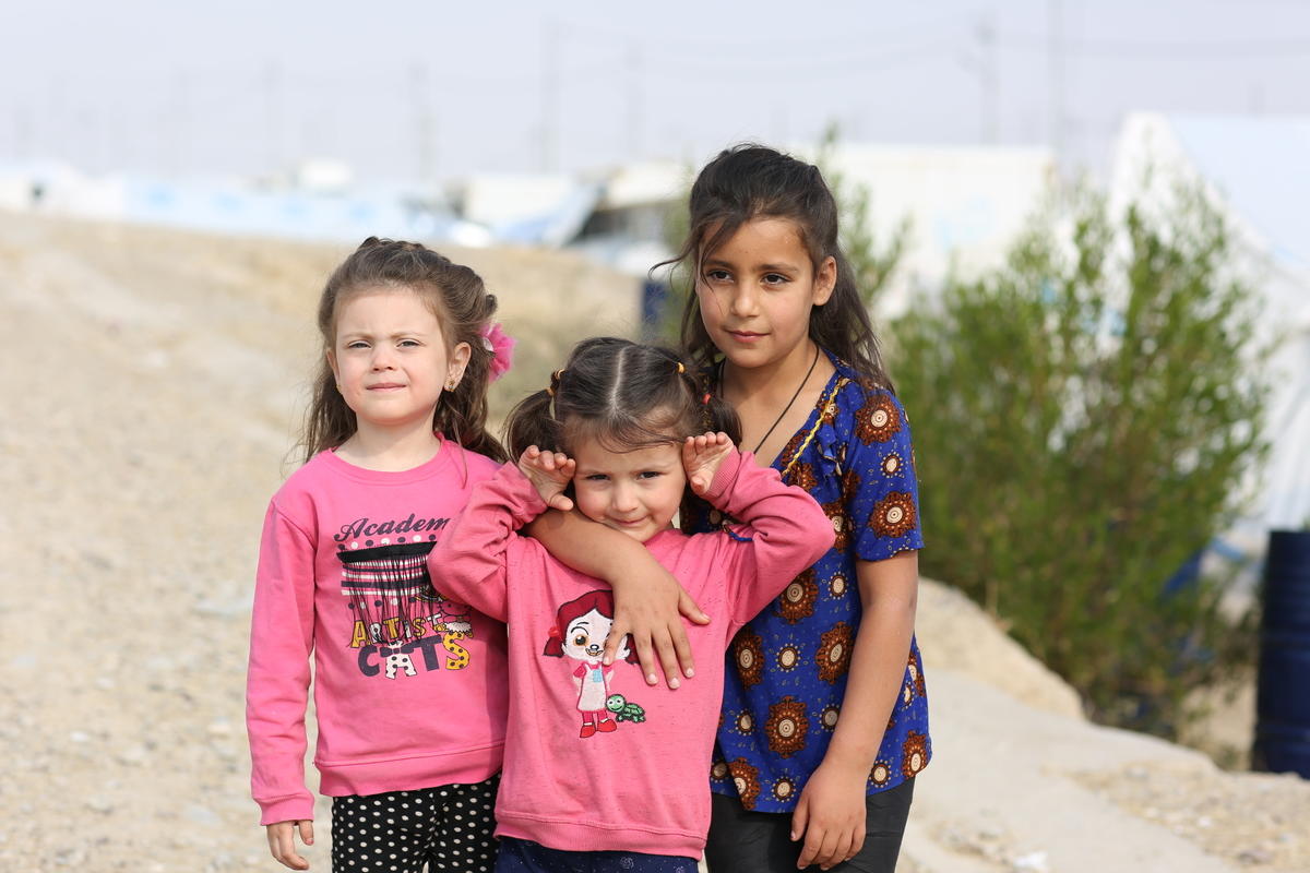Three Syrian girls stand together in a refugee camp, with tents in the background