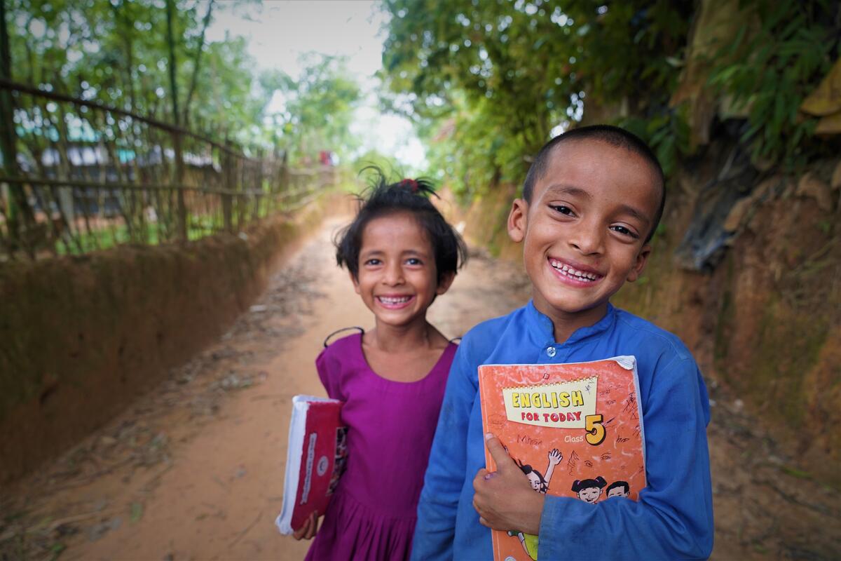 A young boy and girl from Bangladesh stand on road, smiling to camera, while holding school books