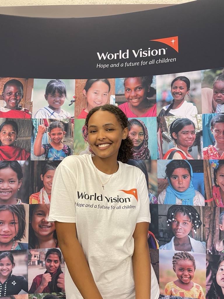Dinna smiles for a photo. She's wearing a World Vision t-shirt and standing in front of a display board featuring the faces of lots of children.