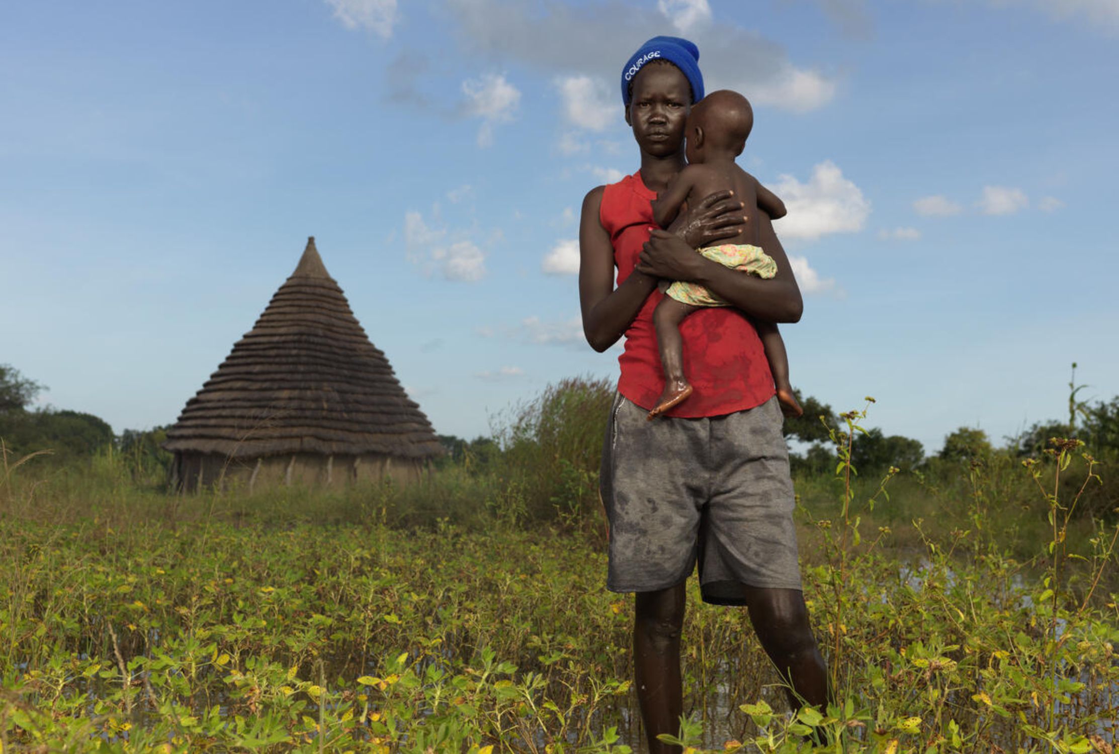 A girl holding her baby, stands in a field nearby a hut