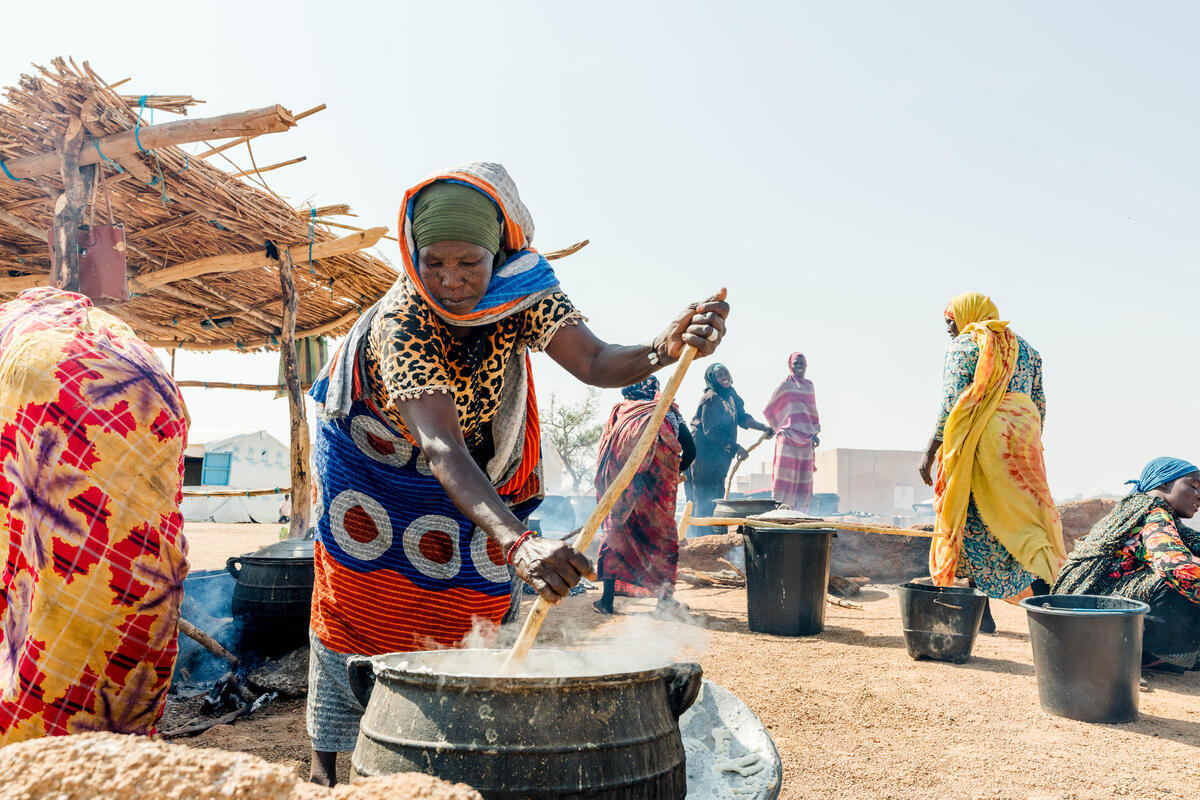 A woman is seen stirring with a spatula over a boiling pot, there are other women in the background