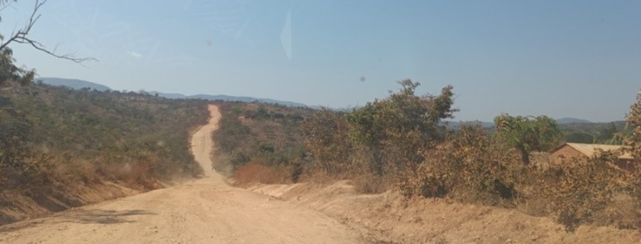 Dusty dirt road with dry vegetation, trees and hills under a clear blue sky