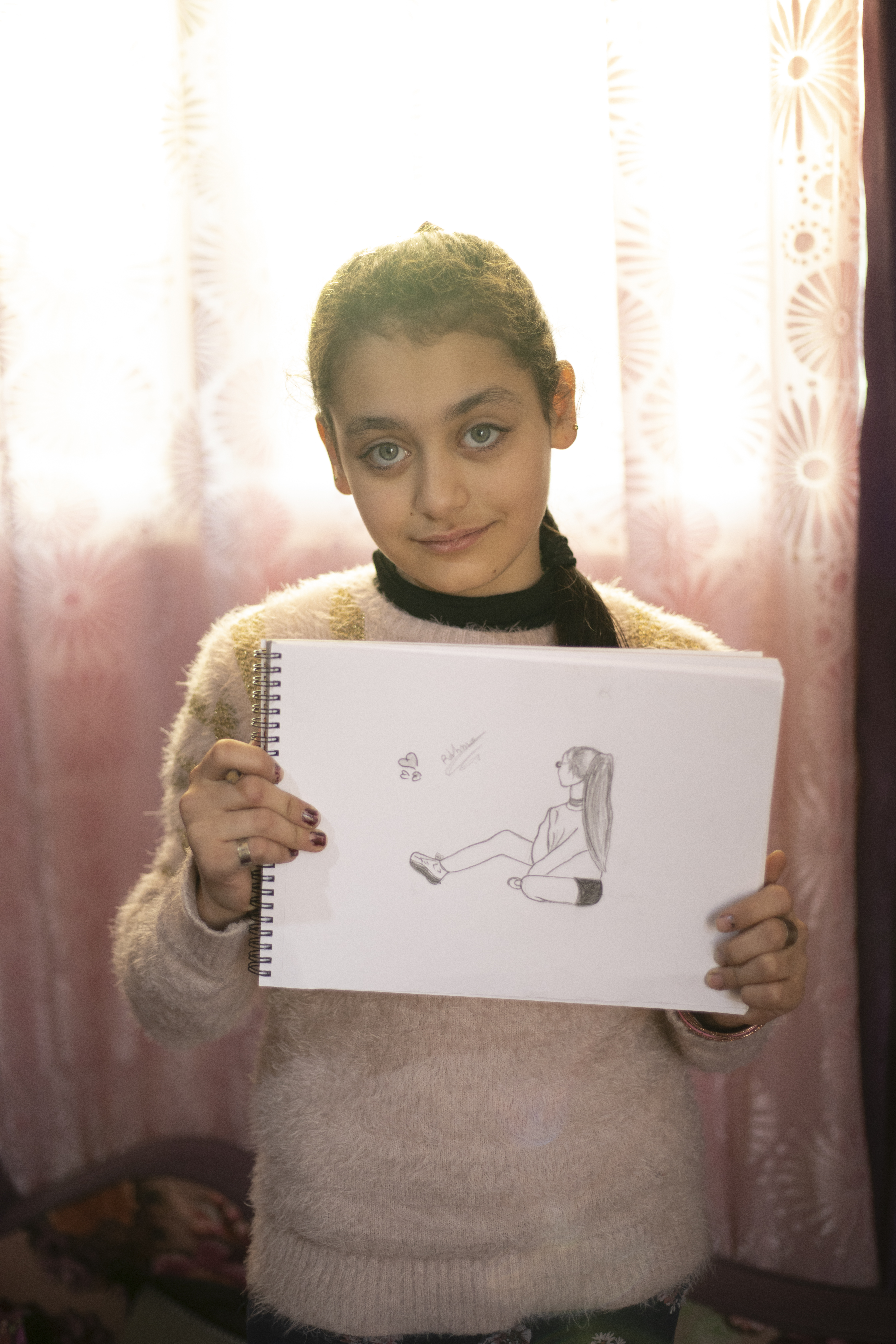 A young girl from Syria, standing indoors, holding one of her drawings featuring a women.