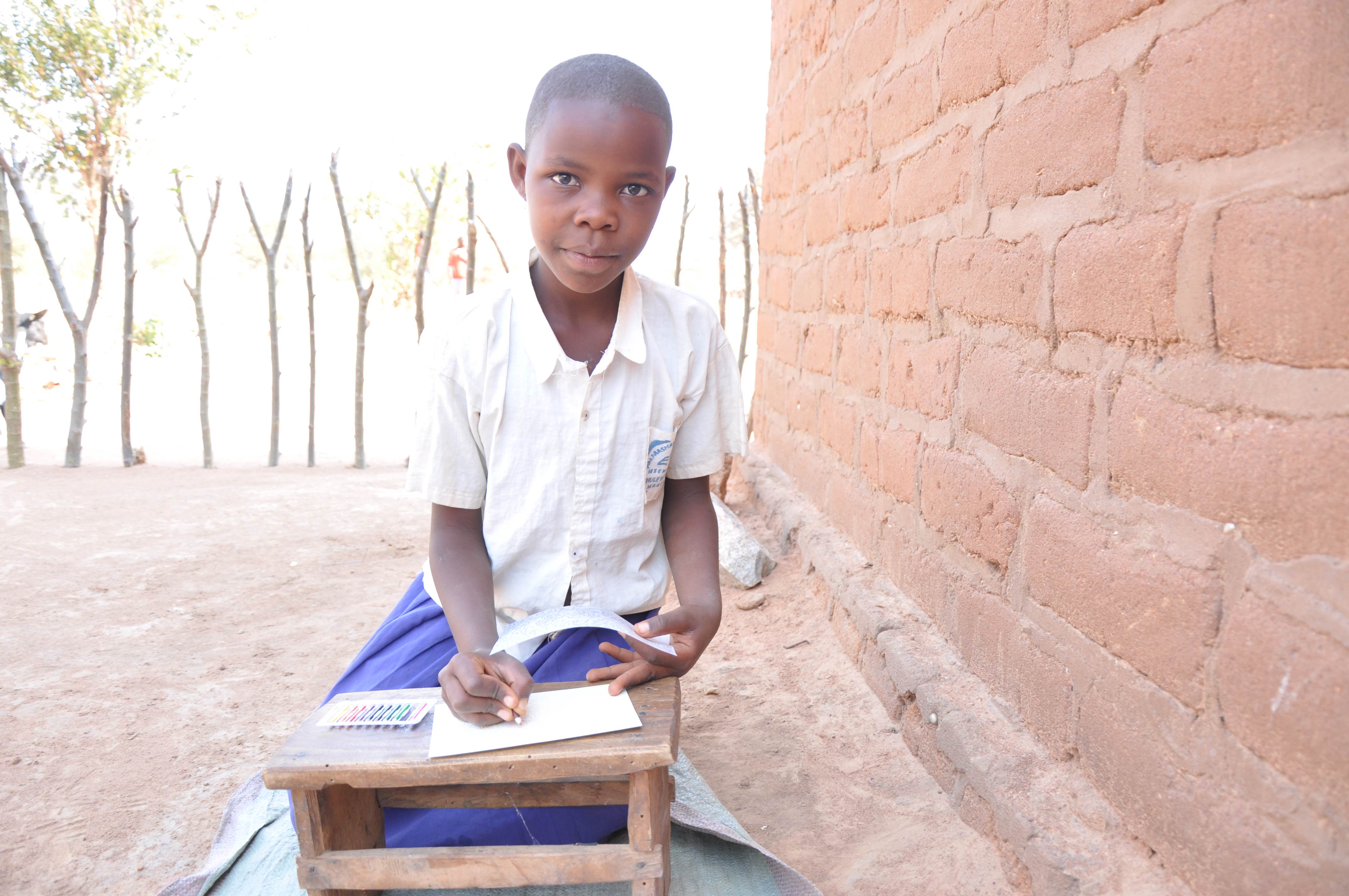 Child from Tanzania smiles at the camera, holding hand out to write on a book, sitting outside