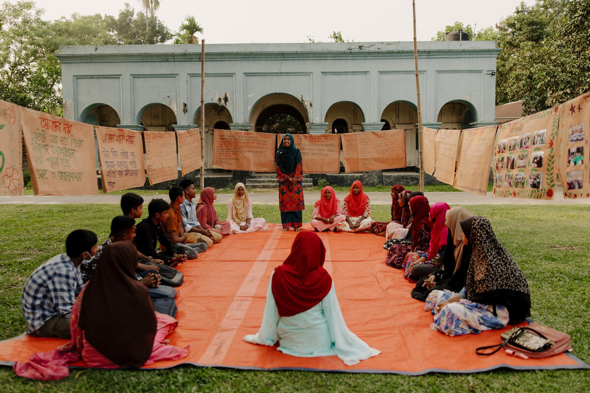  Misu stands and addresses a group of young people seated in a circle outdoors in front of a building in Bangladesh.