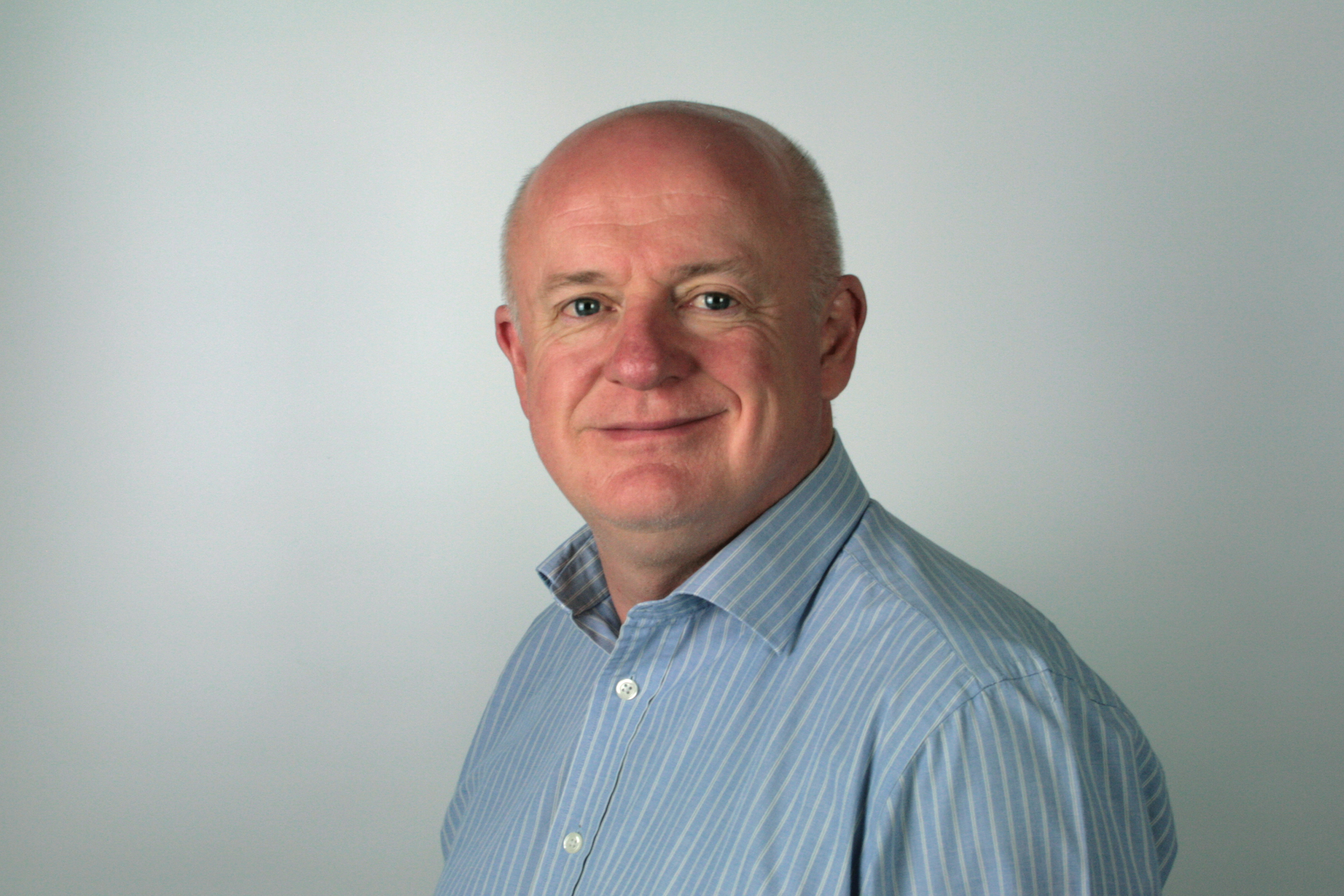 Man in the UK smiles as he wears a blue shirt, against a white wall backdrop