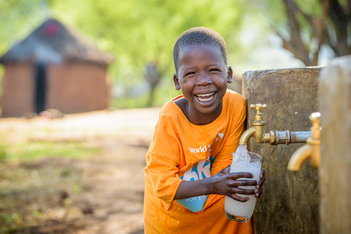 A little girl called Cheru stands in an orange t-shirt smiling by a water point in her village