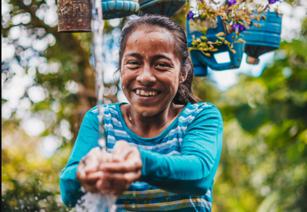 A teenage girl smiling, washing her hands in clean water under an outdoor tap