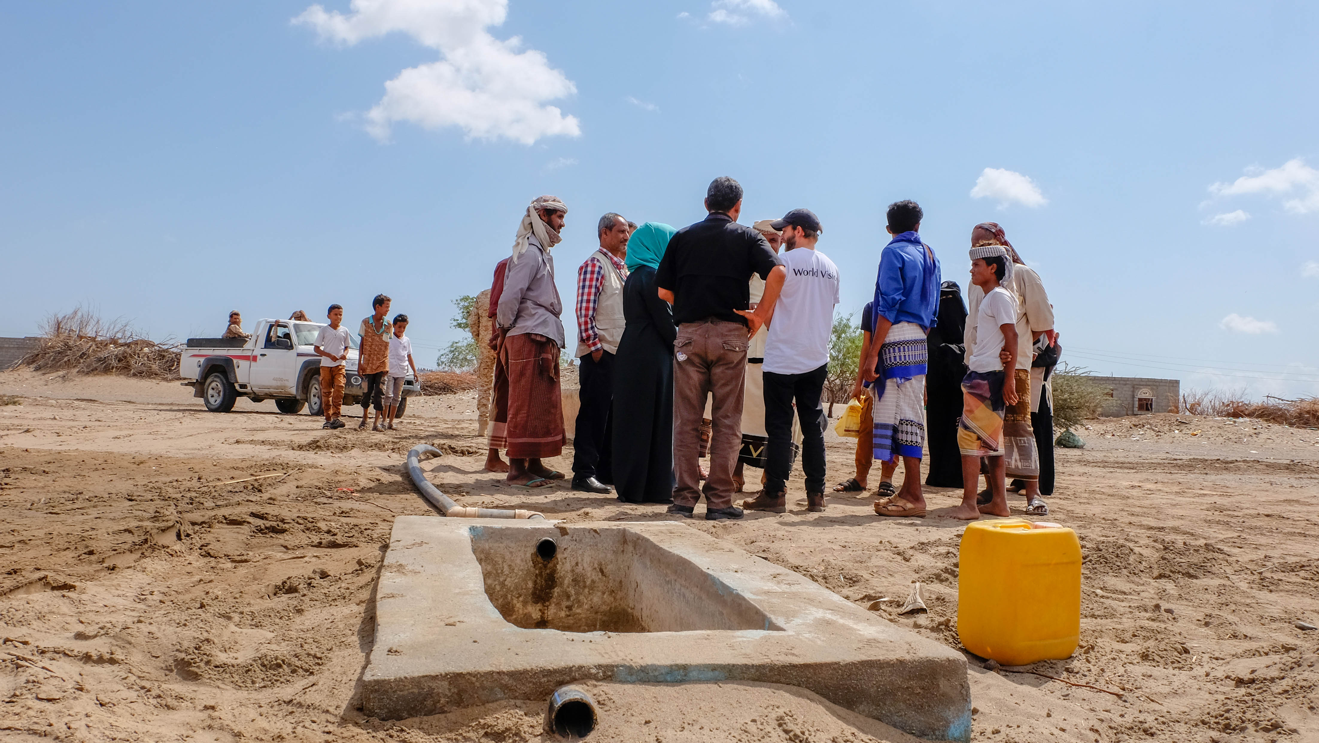 A group of people in Yemen stand in front of a tap in a barren landscape under a blue sky