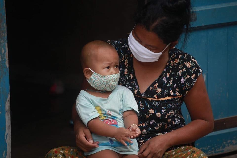Toddler and his mum sit in their doorway in Thailand. They are both wearing masks to protect themselves from coronavirus.
