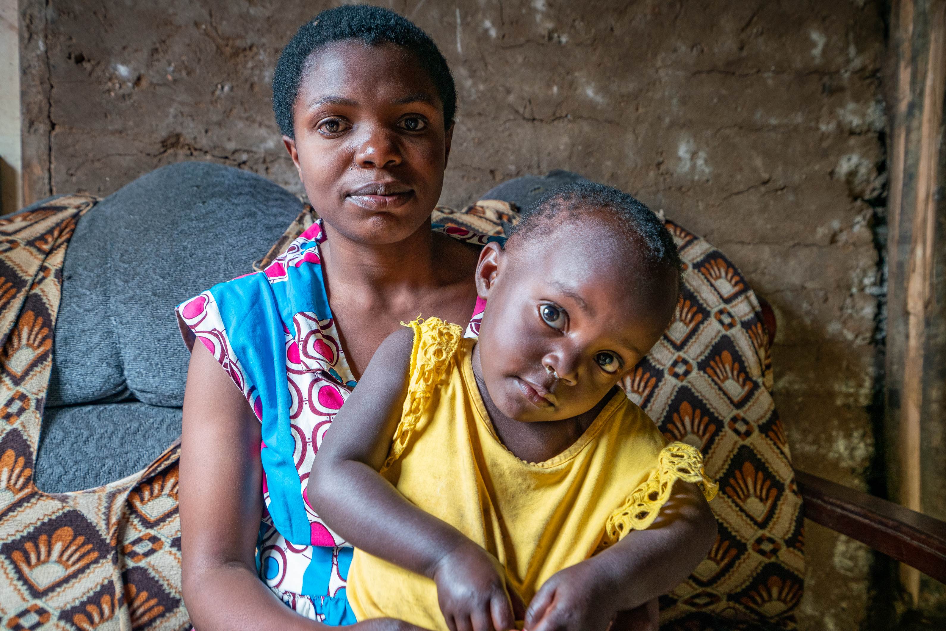 Woman from DRC sits with a child on her lap, looking into the camera against a brown wall