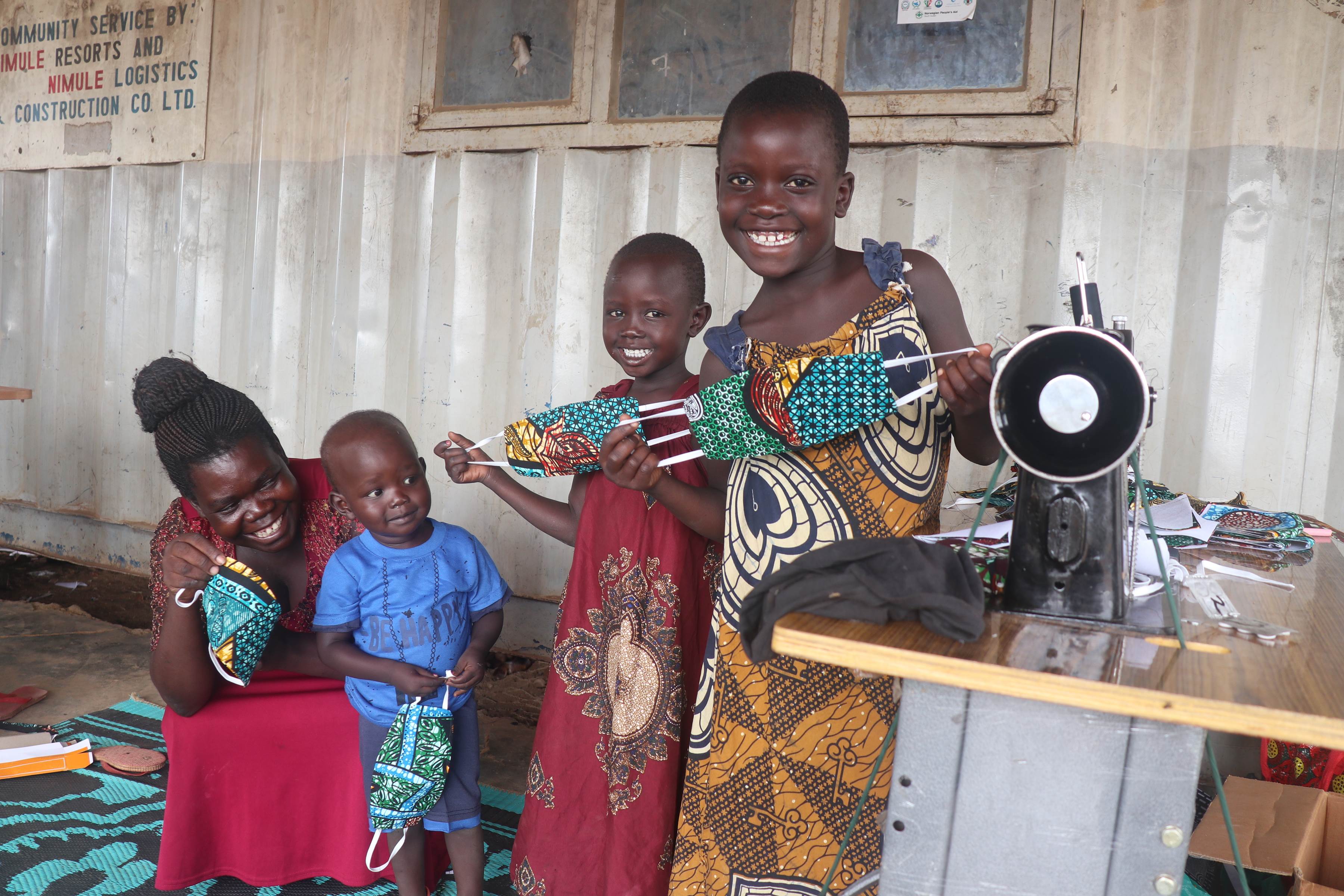 Two women and two children smile and hold out garments, standing next to a sewing machine in South Sudan