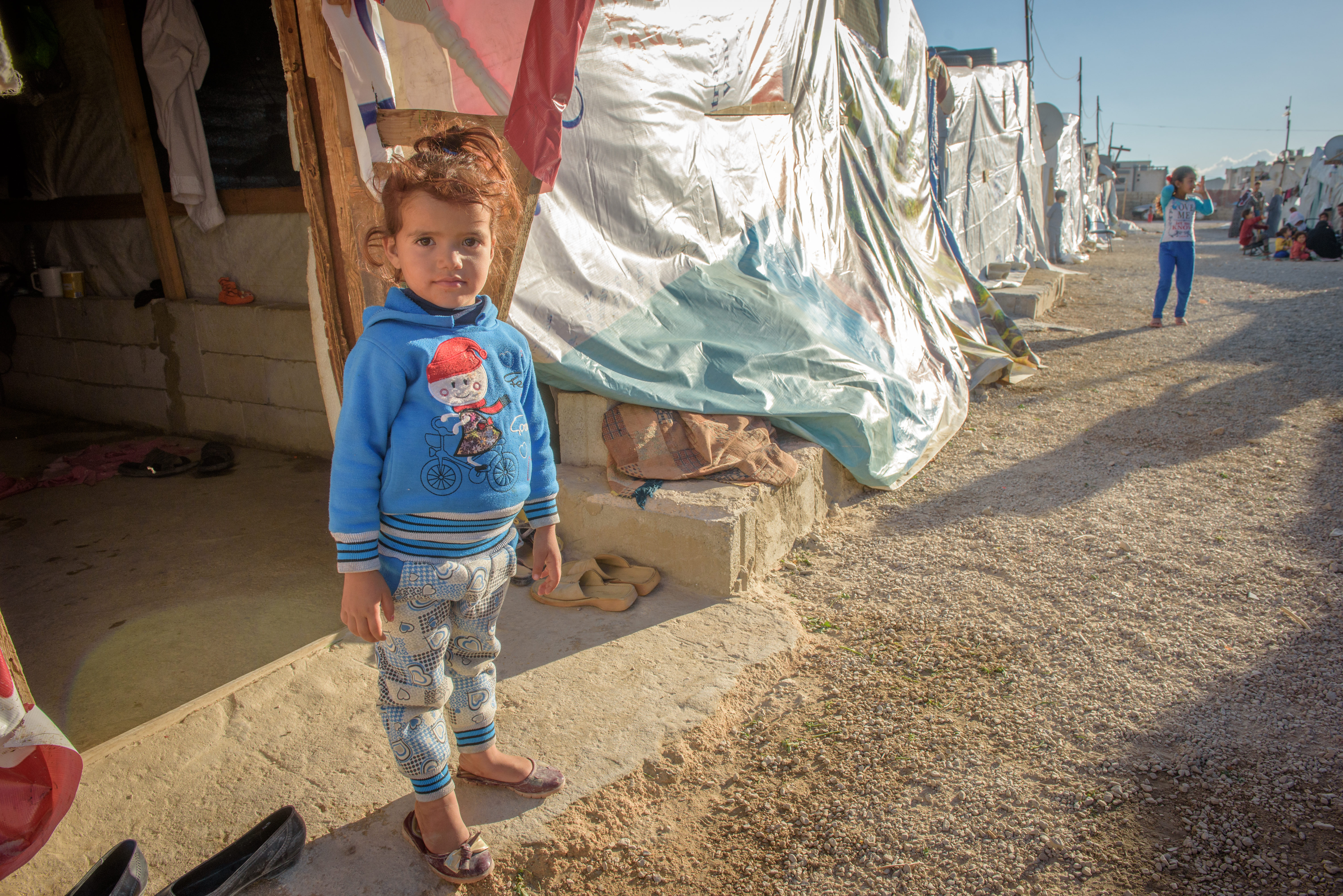 Girl stands outside a tent in a refugee camp in Lebanon, next to a line of more tents